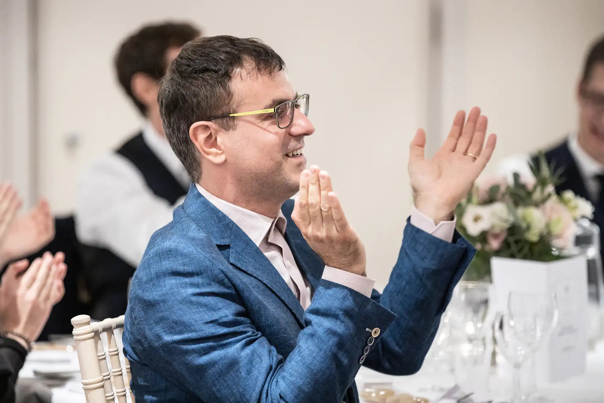 A man in a blue blazer is seated at a formal event table, clapping and smiling, with other people clapping in the background.