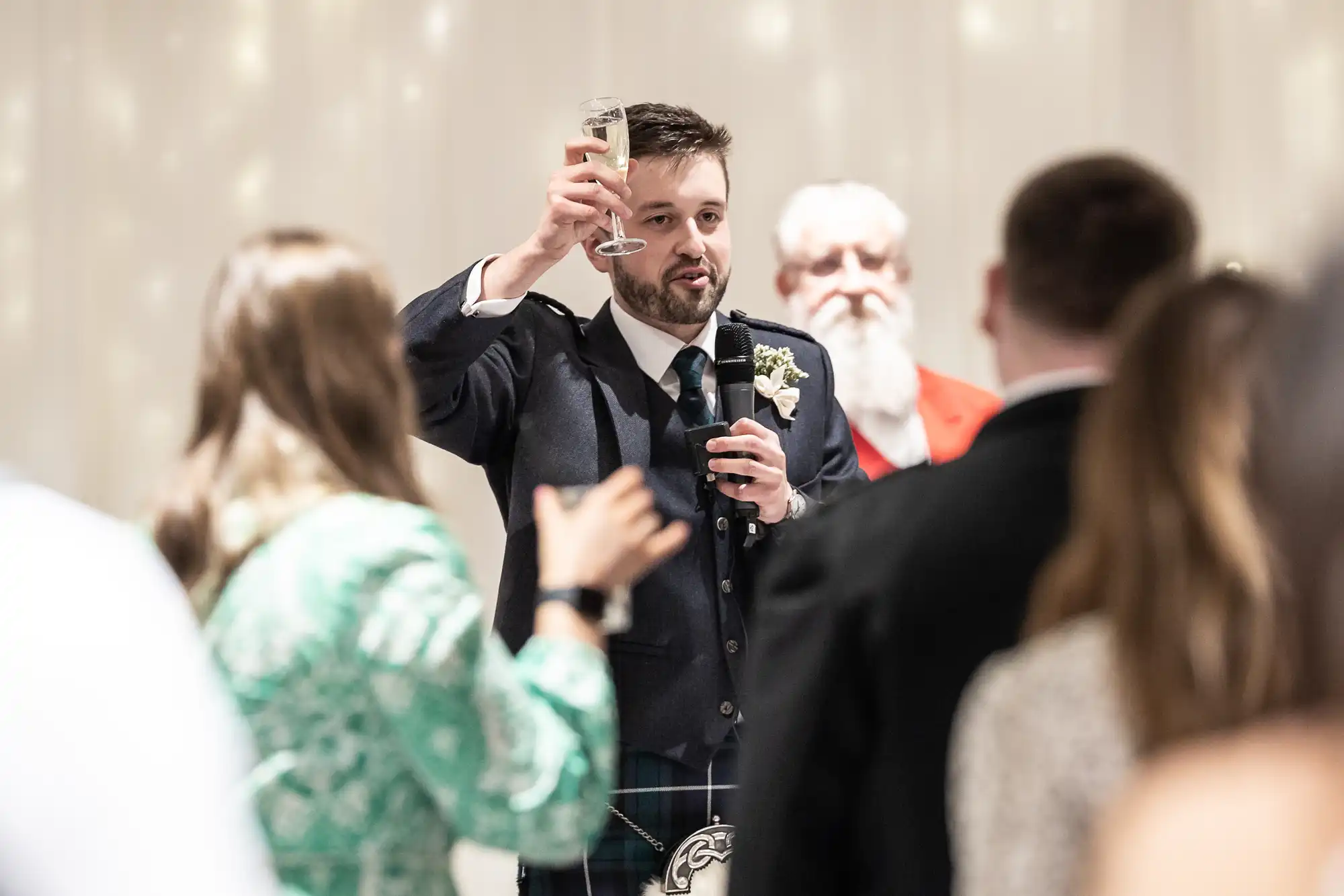 A man in formal attire raises a glass while speaking into a microphone. Several people stand around him, including an older man in a red jacket.