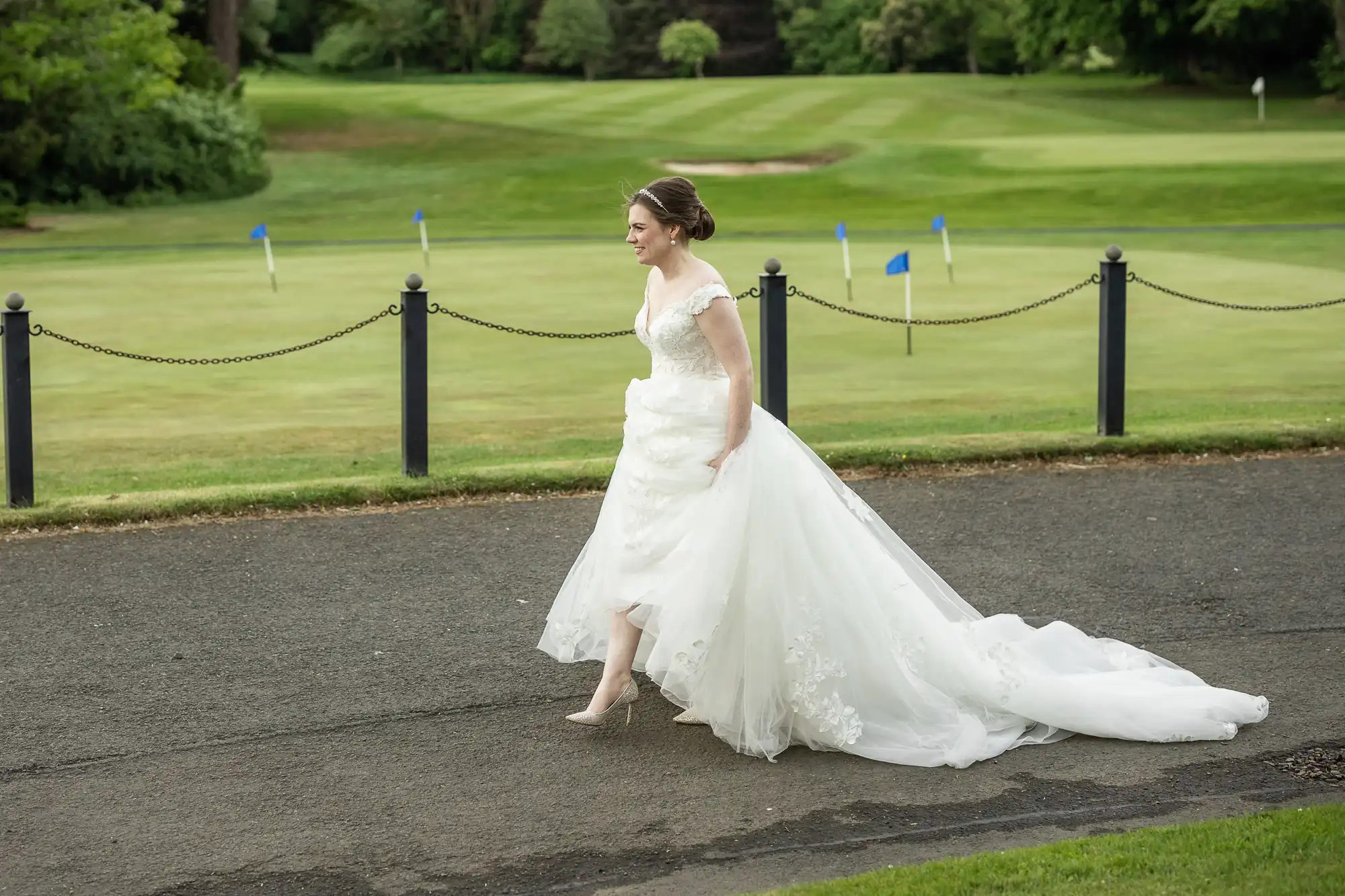 A woman in a white wedding dress walks on a paved path next to a grass field with blue flags.