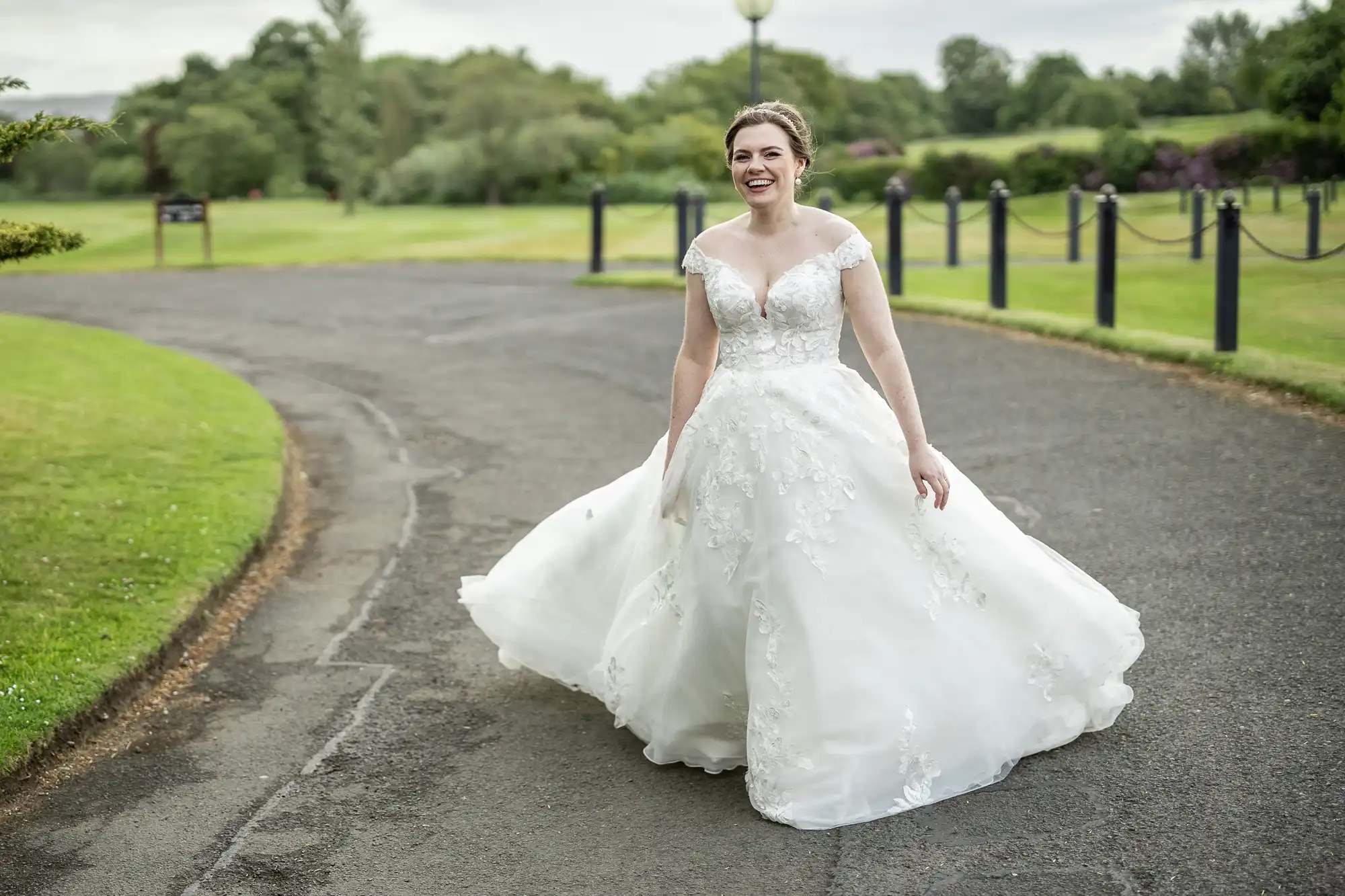 A woman in a white wedding gown smiles while walking on a paved path in a park with manicured lawns and trees in the background.