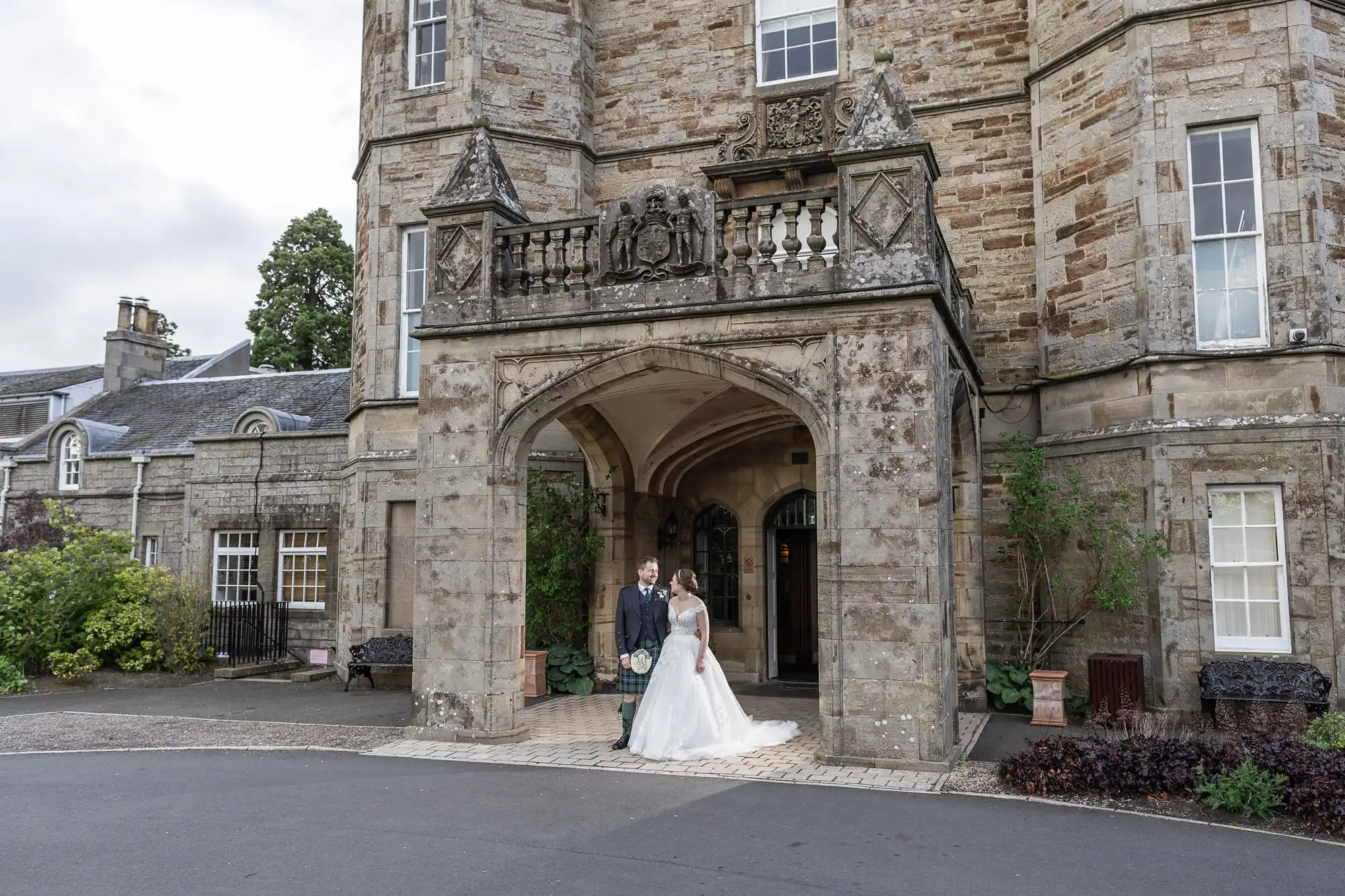 A couple in wedding attire stands in front of a large, historic stone building with an ornate arched entrance and detailed architectural features.