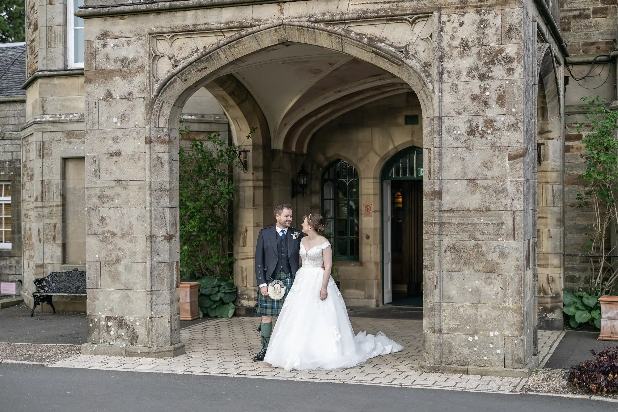 A bride and groom stand together outside an arched entrance of a stone building, both looking at each other. The groom is wearing a traditional kilt, and the bride is in a white wedding dress.