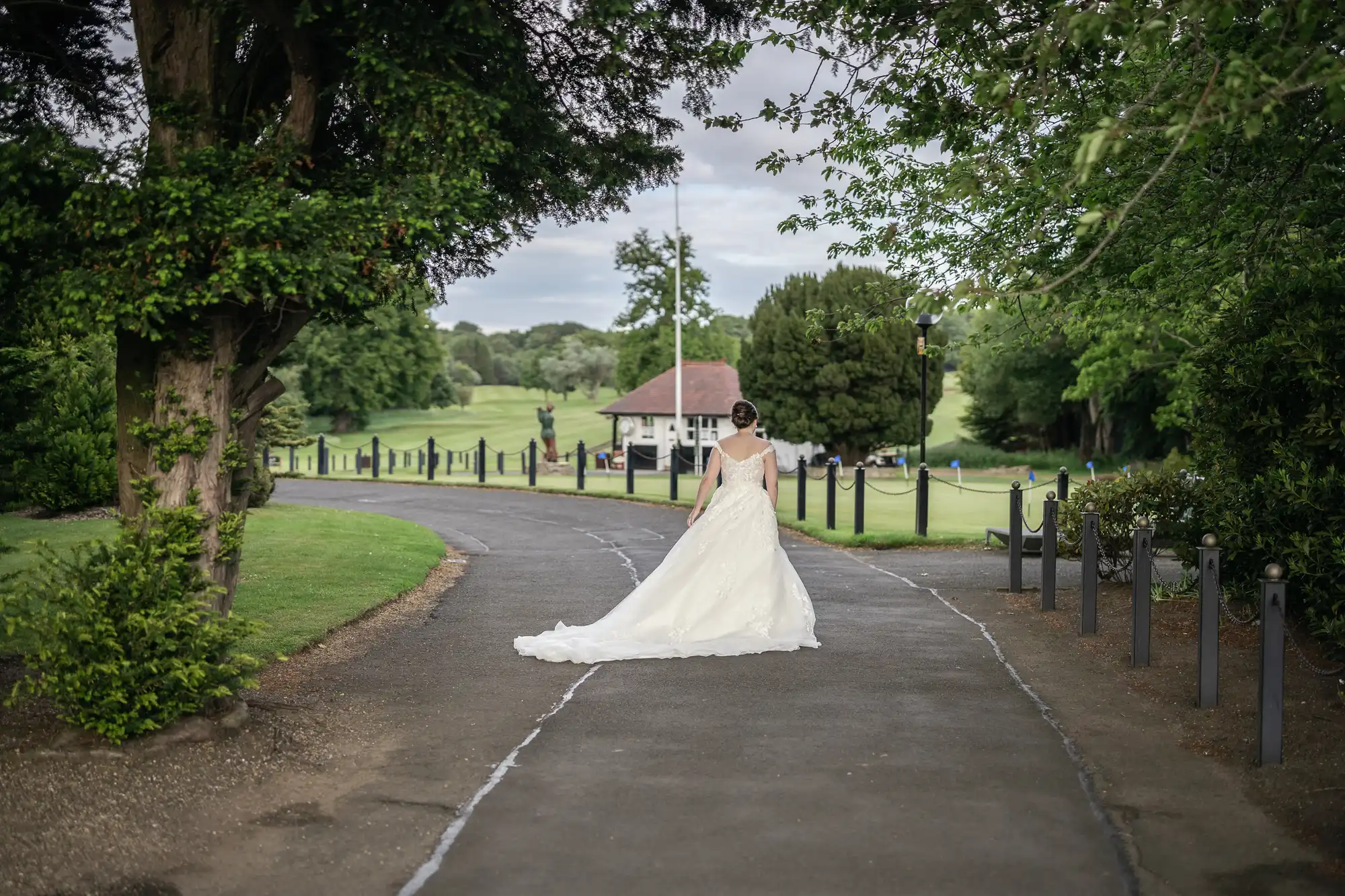 A bride in a white wedding dress walks alone down a paved path in a park with trees and a small building in the background.