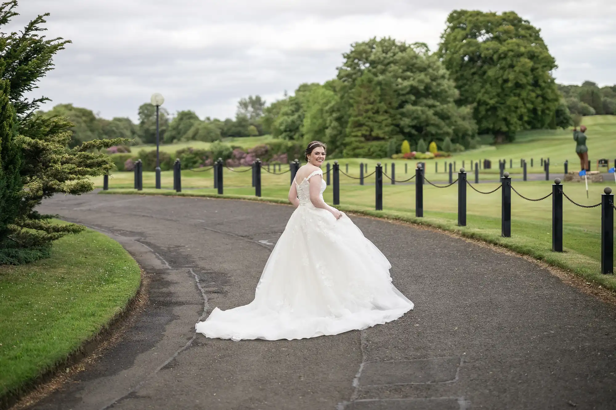 A bride in a white wedding dress walks down a paved path lined with black posts and chains, with manicured lawns and trees in the background.