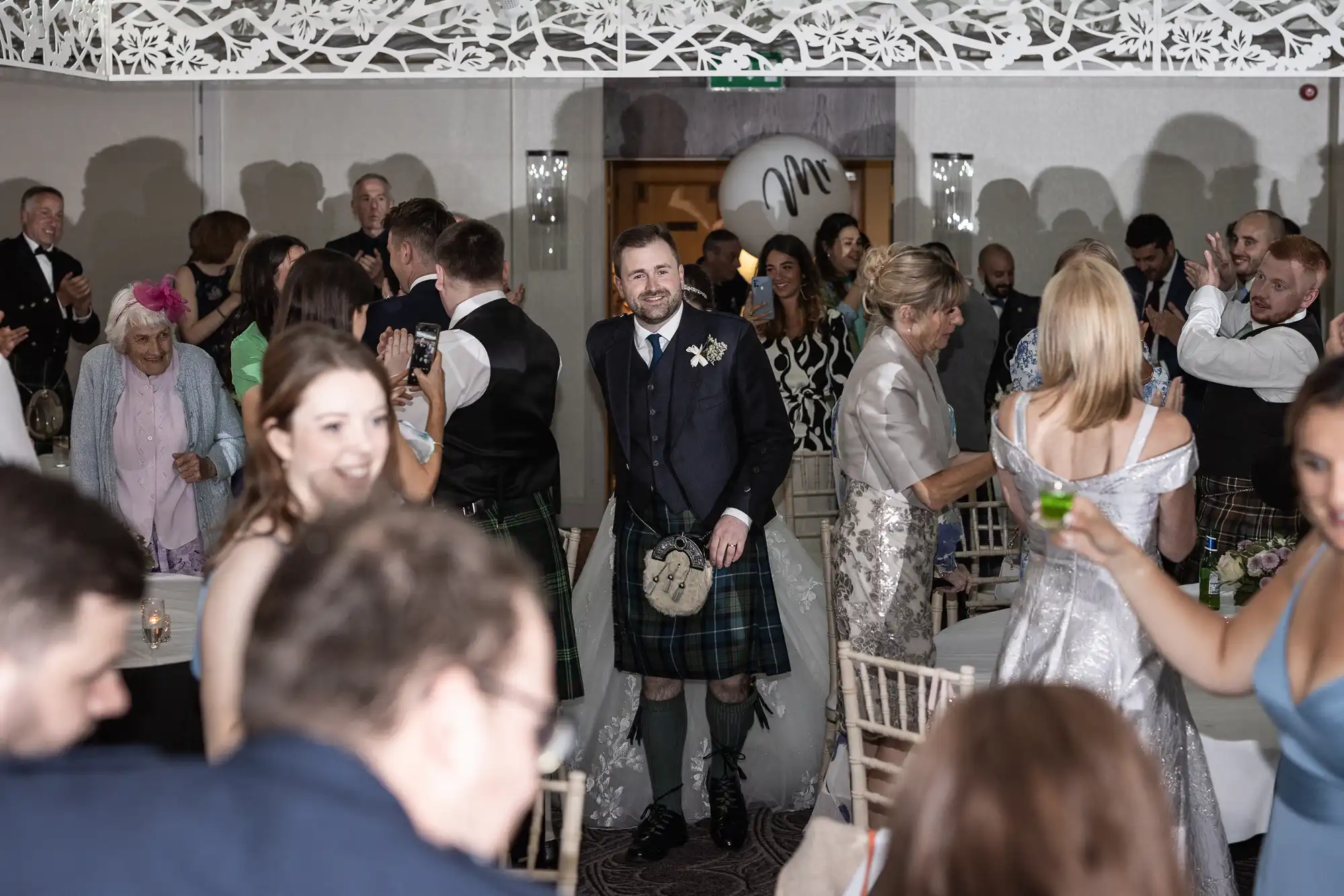 A man in a kilt walks through a crowd of people at a wedding reception. Guests, dressed in formal attire, are clapping and smiling.