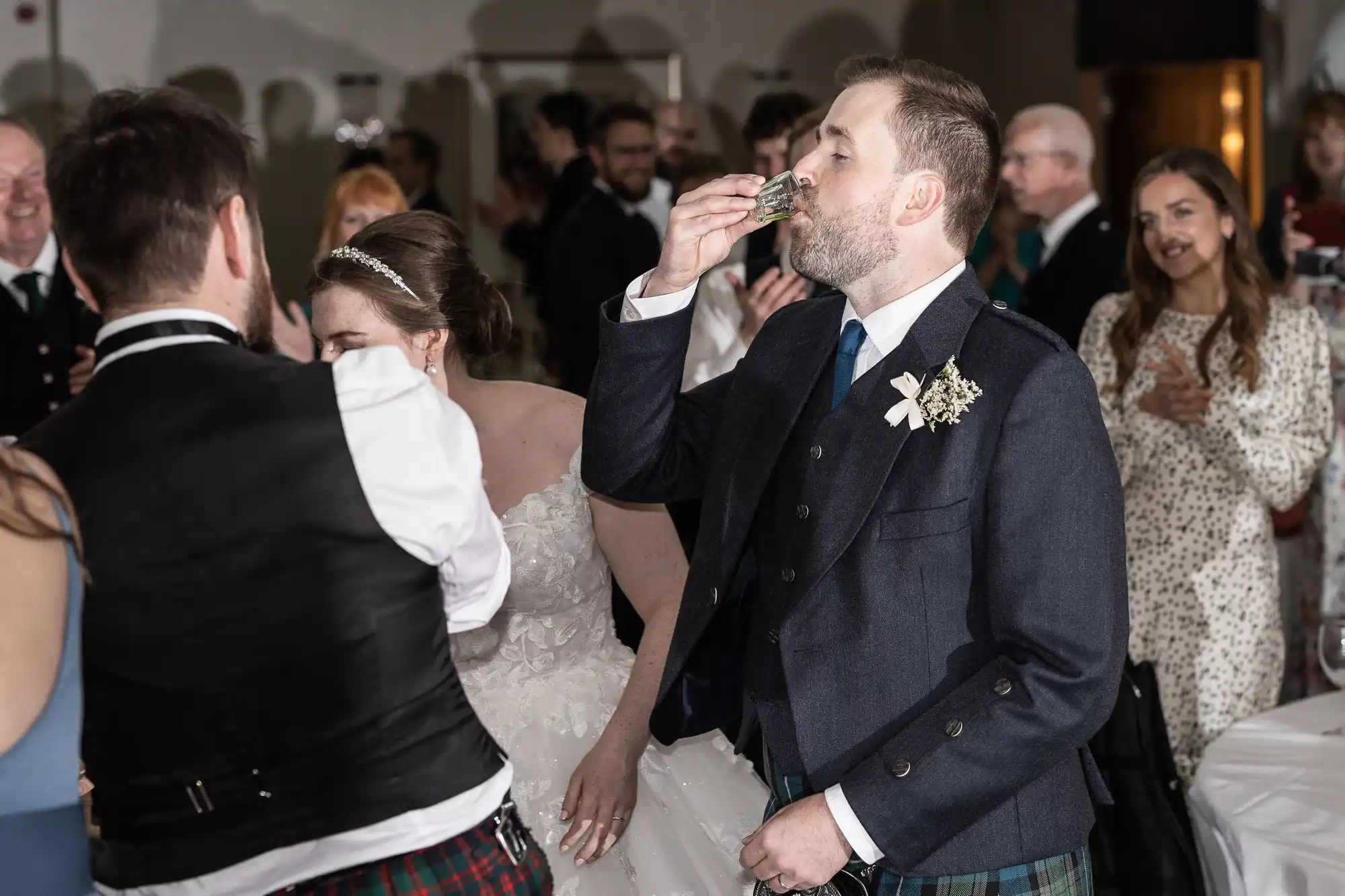 A groom in traditional attire drinks from a small cup at a wedding reception as guests look on and applaud.