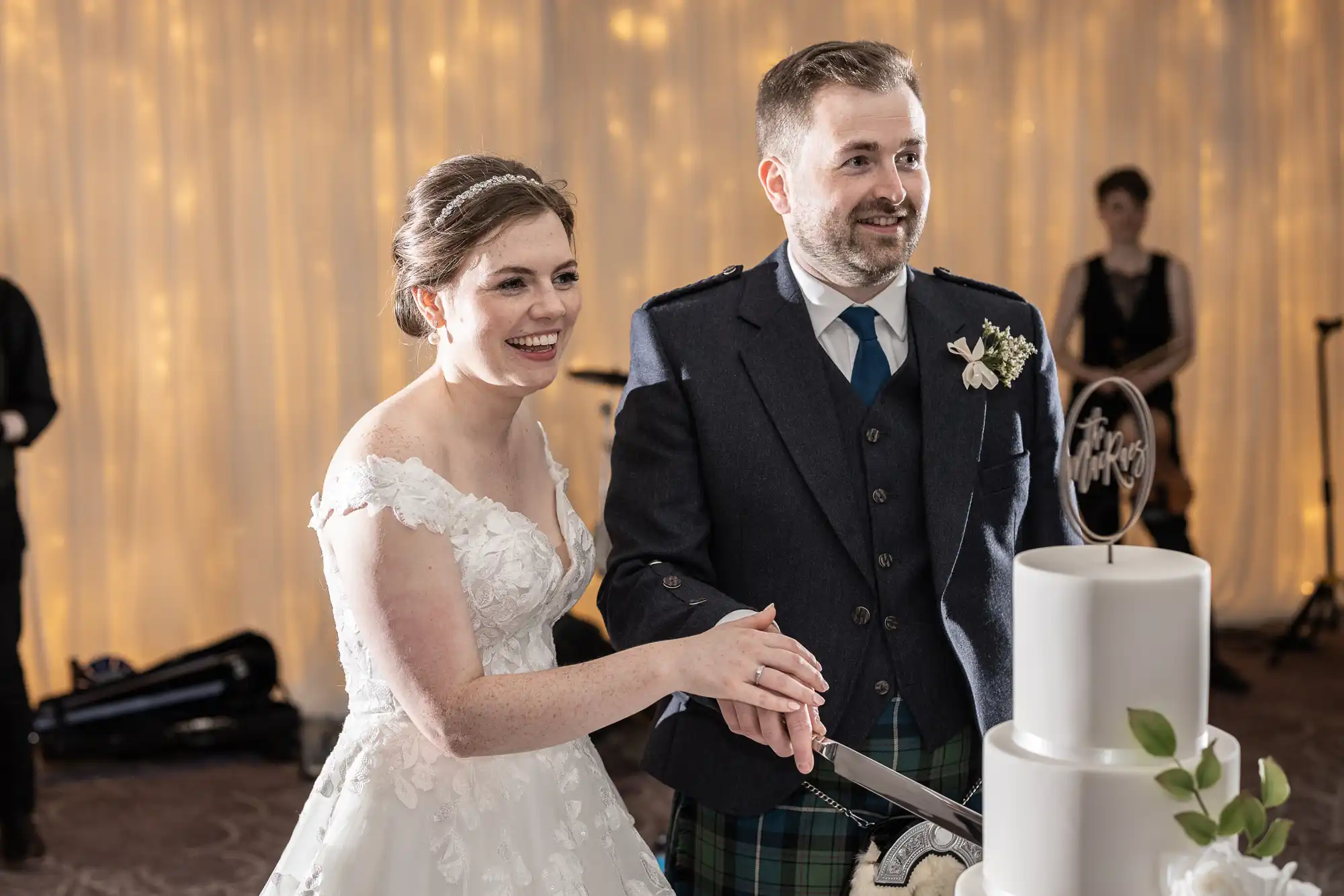 A bride and groom dressed in a wedding gown and Scottish attire, respectively, smile as they cut a white, three-tiered wedding cake.