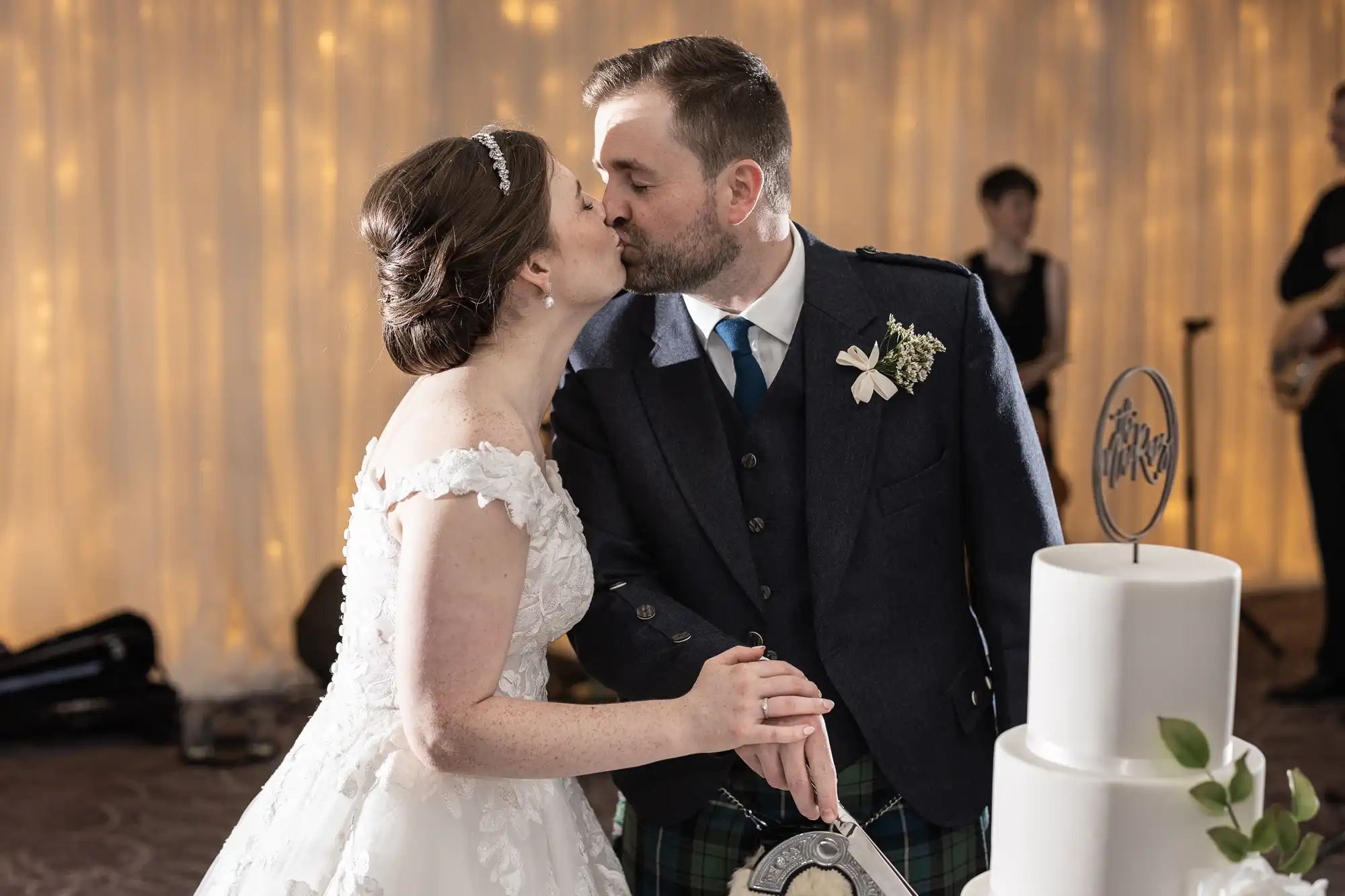 A bride and groom kiss while cutting a tiered wedding cake. The bride wears a white dress, and the groom is in a suit with a plaid kilt.