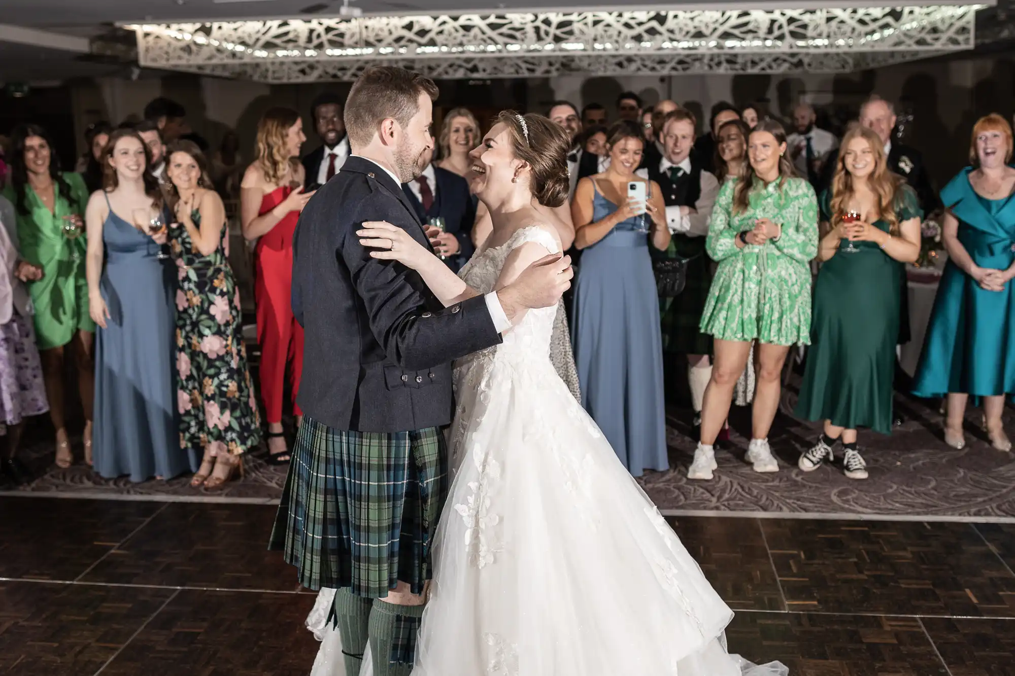 A bride and groom share their first dance at their wedding, surrounded by smiling guests who are watching and taking photos.