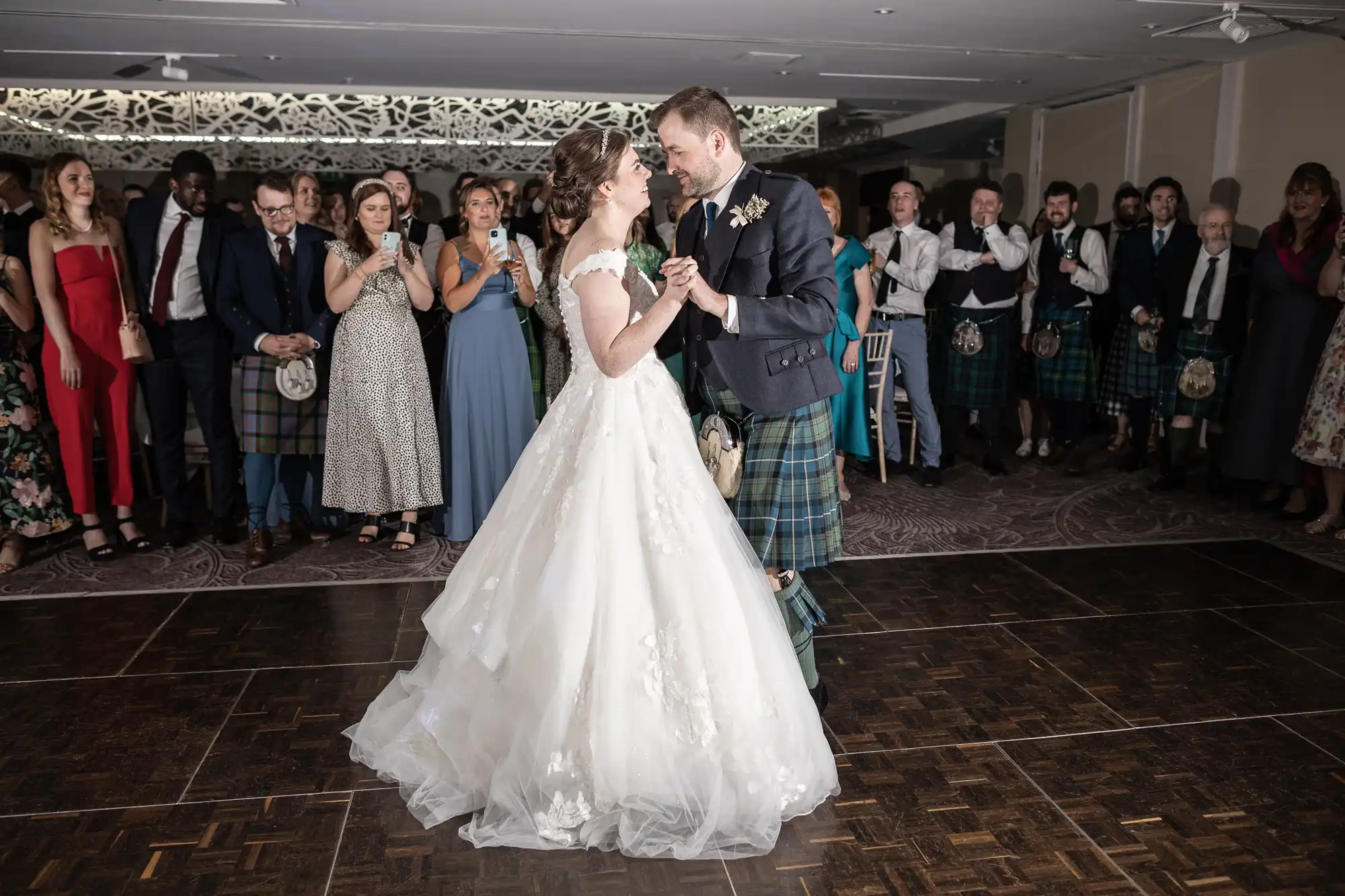 A bride and groom dance in the center of a room surrounded by guests watching and clapping. The bride wears a white gown, and the groom wears a kilt with a dark jacket.