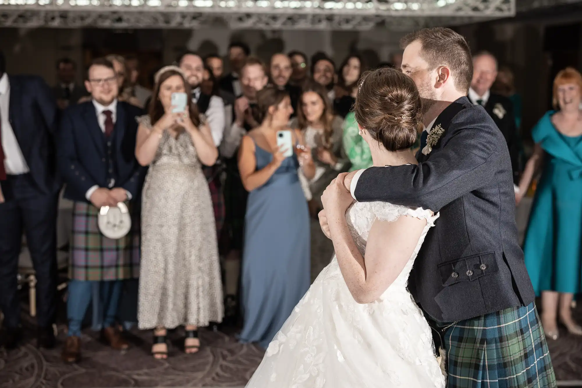 A couple dances at a wedding reception while guests in formal attire, including kilts, watch and take photos.