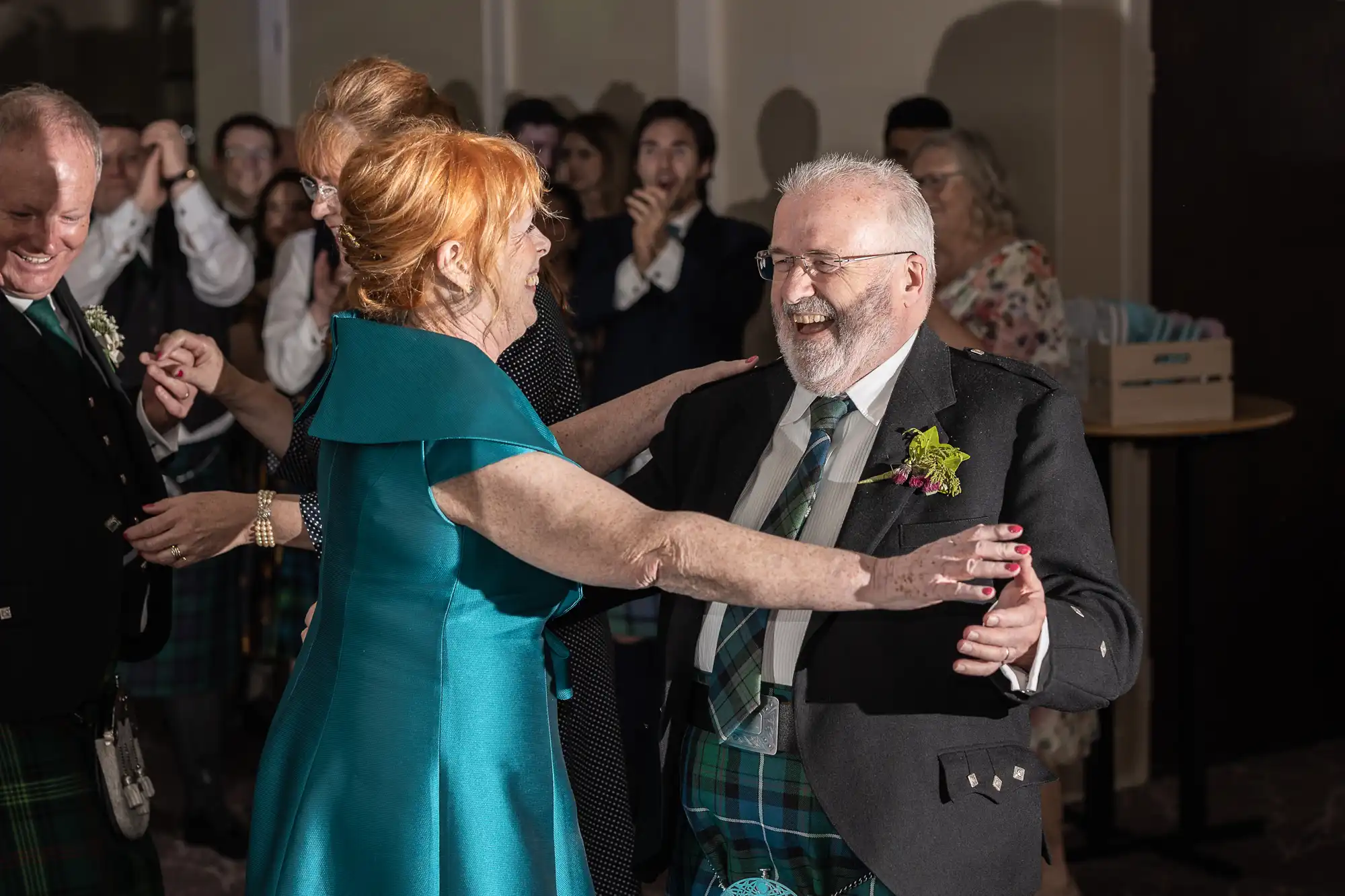 A woman in a teal dress and a man in a suit with a tartan tie joyfully dance together at a celebration. Other people in the background also appear to be enjoying the event.