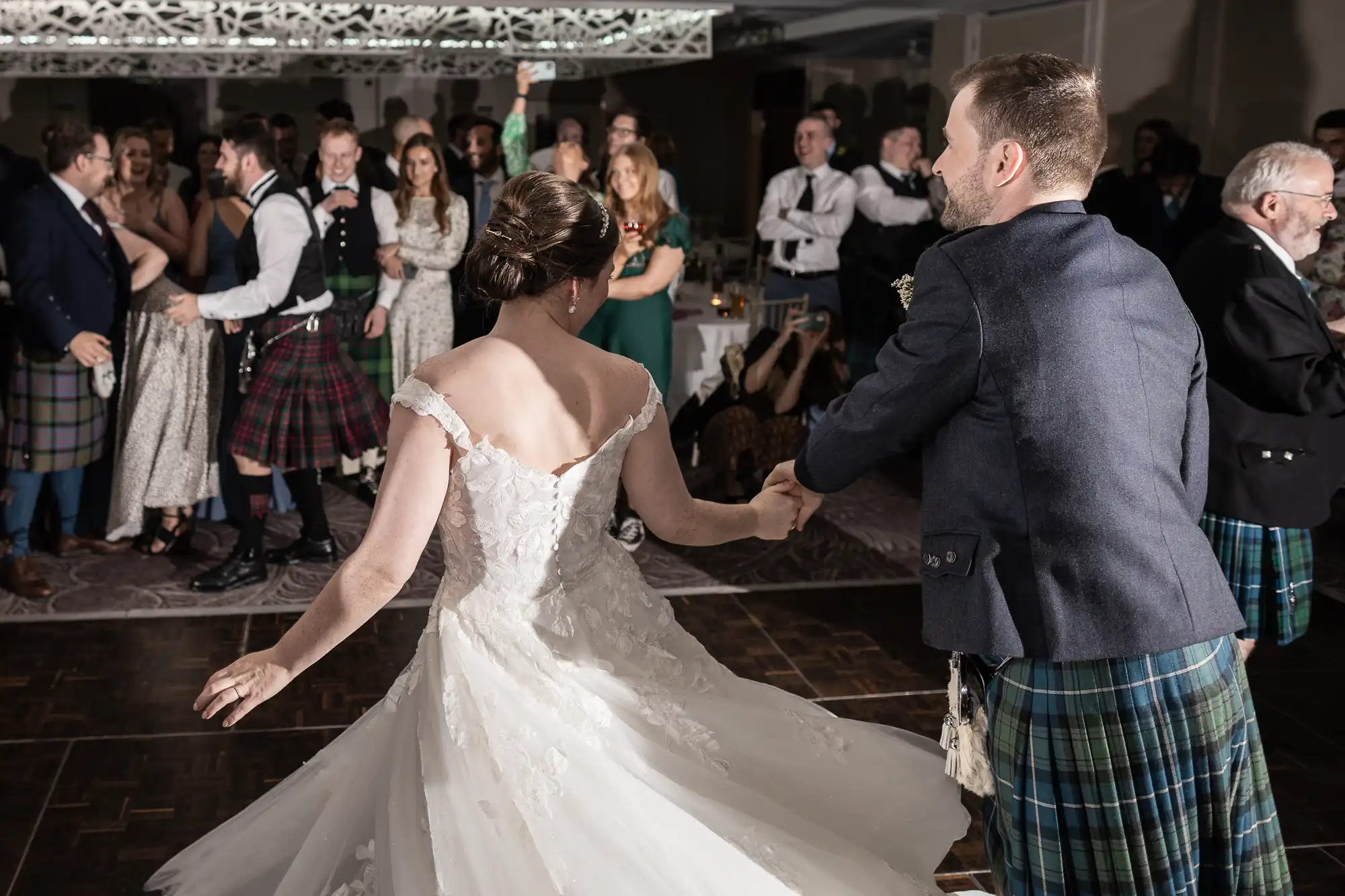 A couple, dressed in a wedding gown and traditional Scottish attire, dance together while guests in formal attire look on and celebrate.