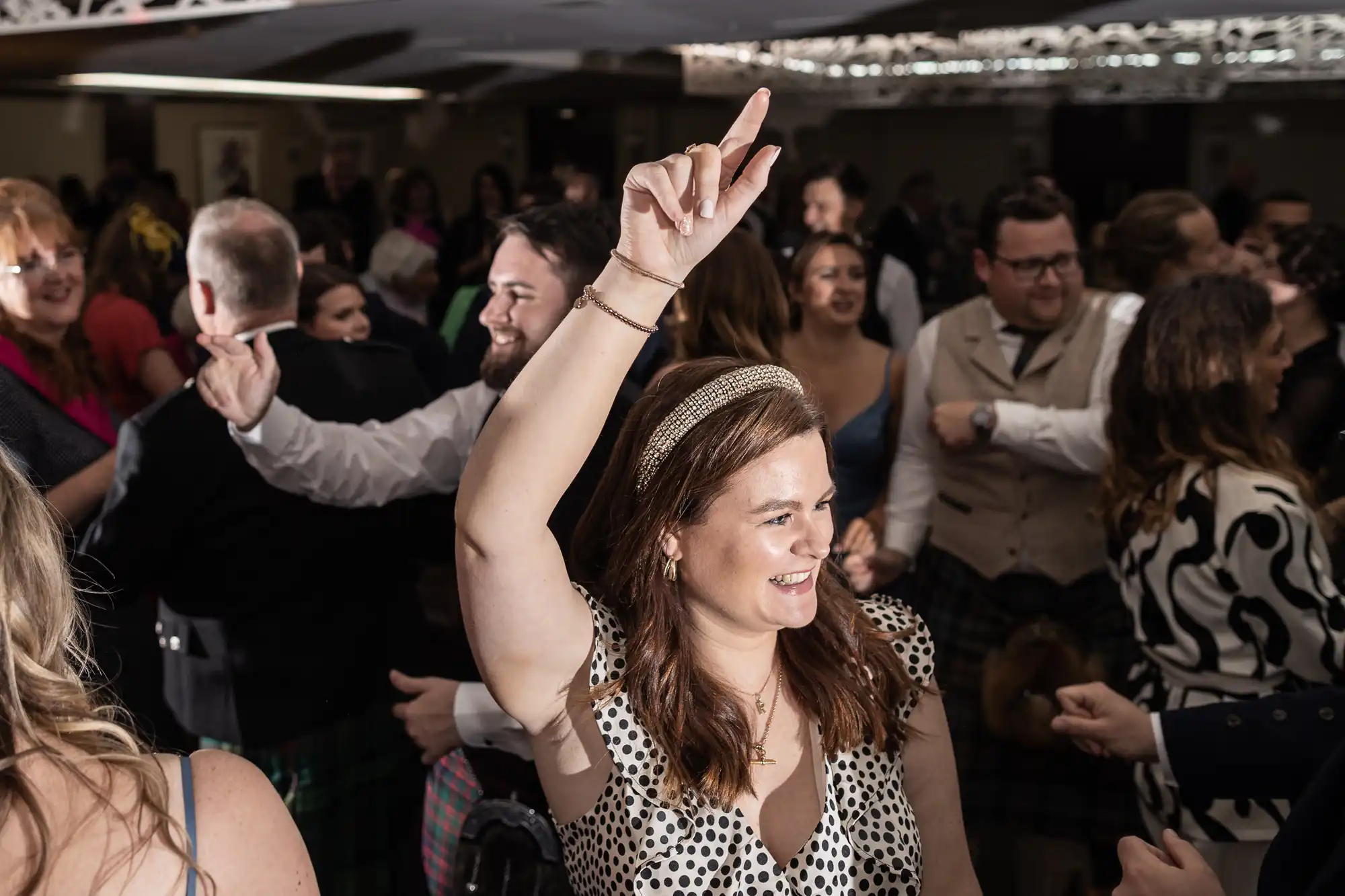 A woman in a polka dot dress dances with one arm raised amidst a crowd of people in formal attire at an indoor event.