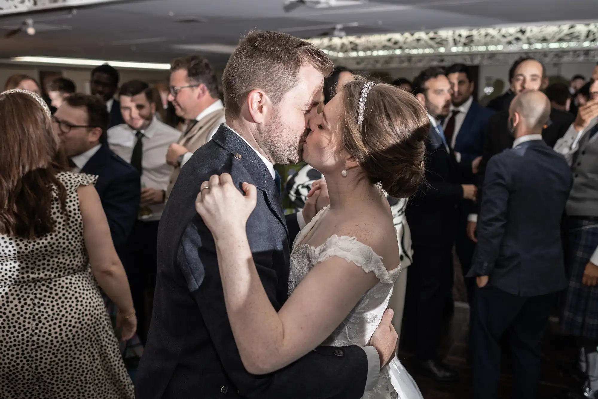 A couple in wedding attire shares a kiss on the dance floor, surrounded by guests dressed in formal wear.