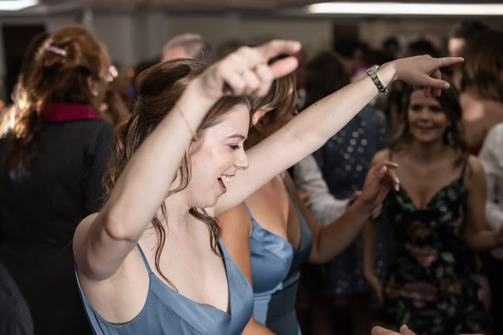 A woman in a blue dress dances with raised arms at a crowded indoor event. People in the background also appear to be dancing and socializing.