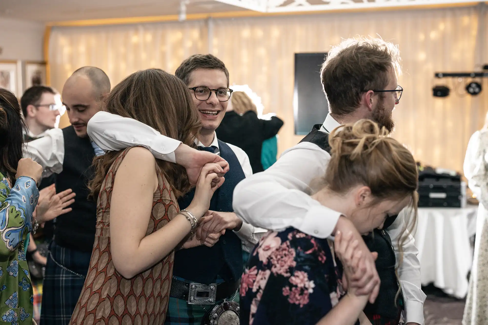 A group of people are dancing closely together at an indoor event, wearing formal attire. One man is smiling directly at the camera amidst the crowd.