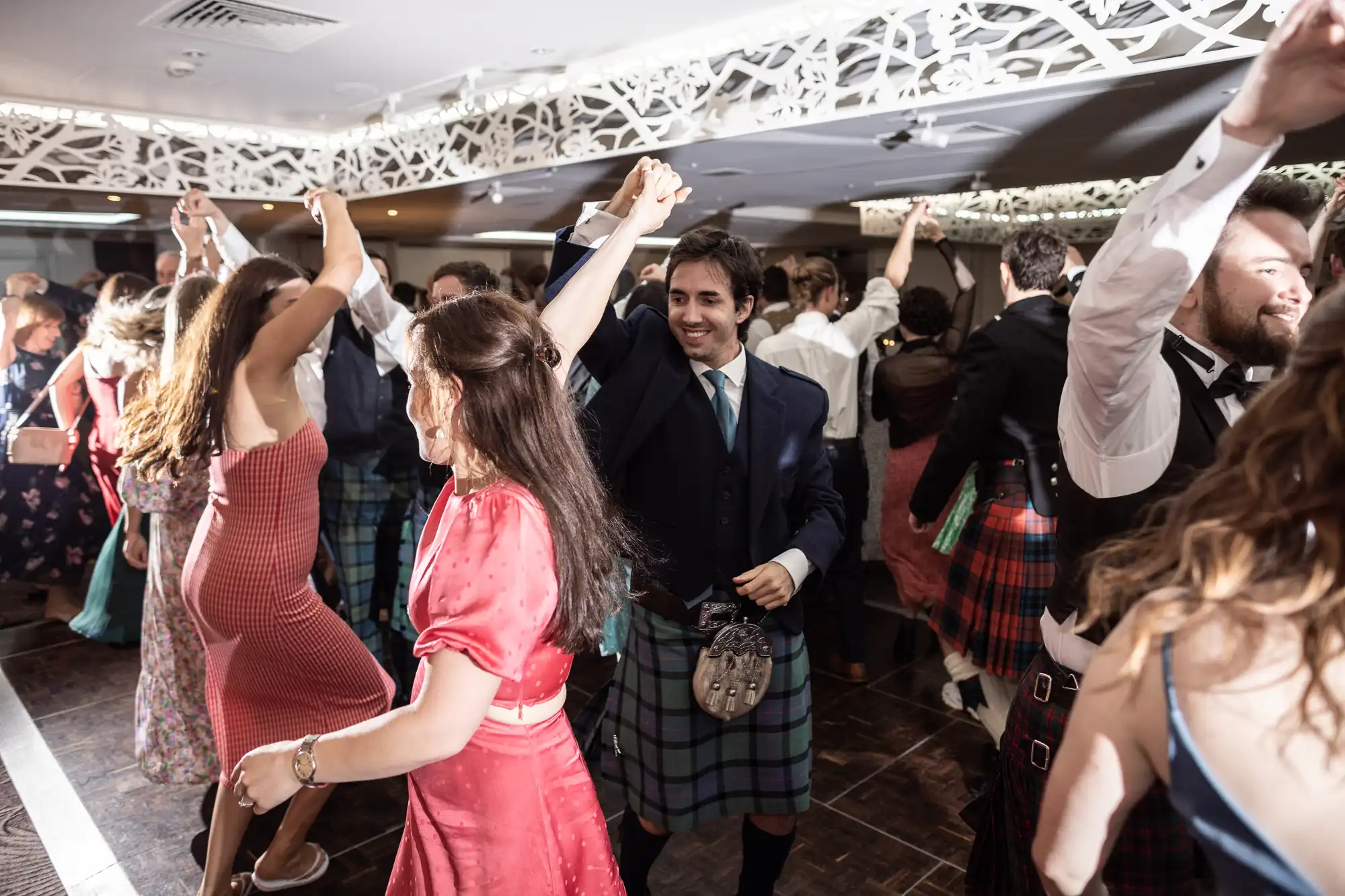 Group of people dressed in festive attire, including kilts and dresses, dancing in a well-lit indoor venue.