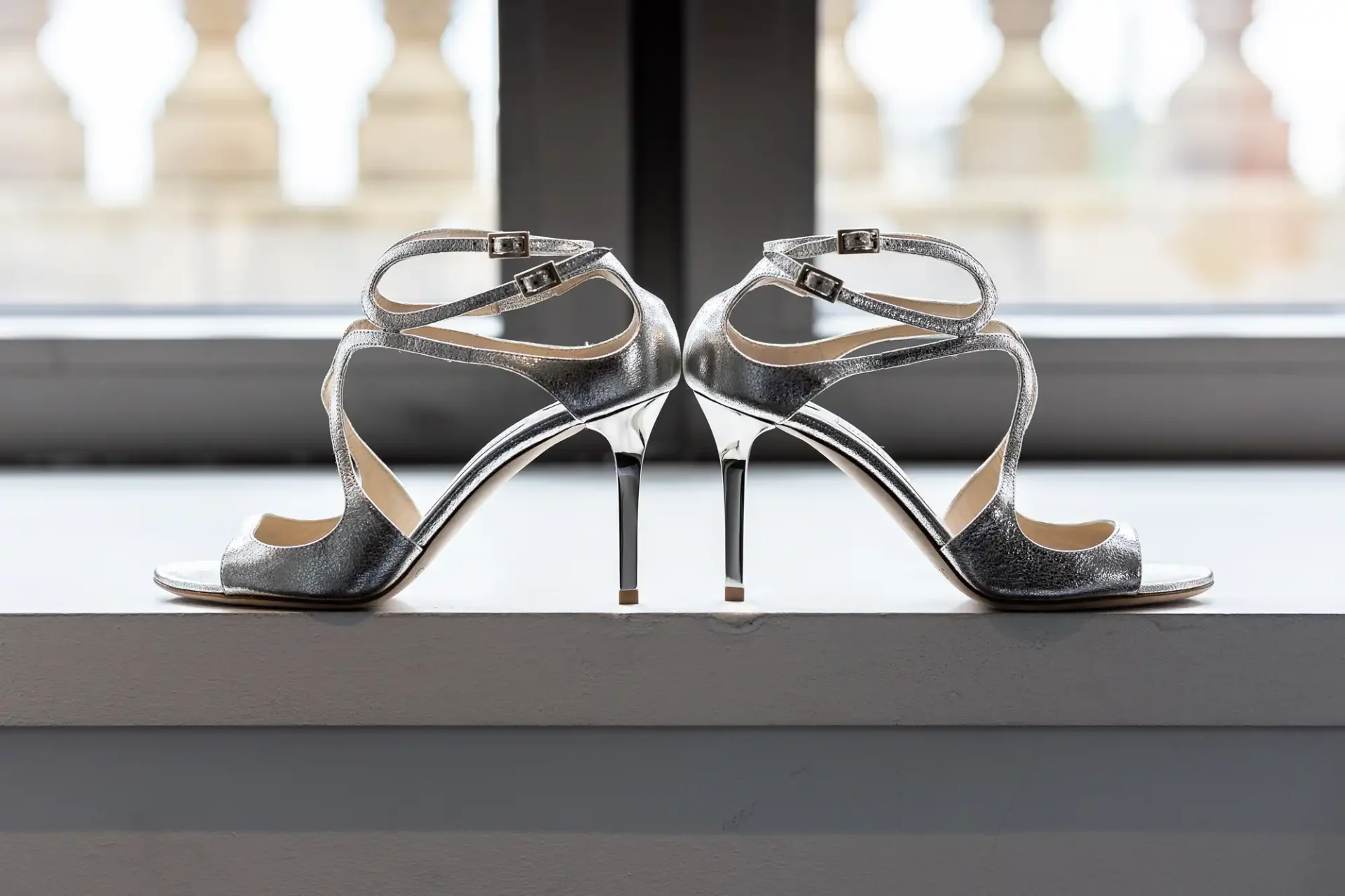 A pair of high-heeled, glittery silver sandals with ankle straps, displayed against a window with blurred background.