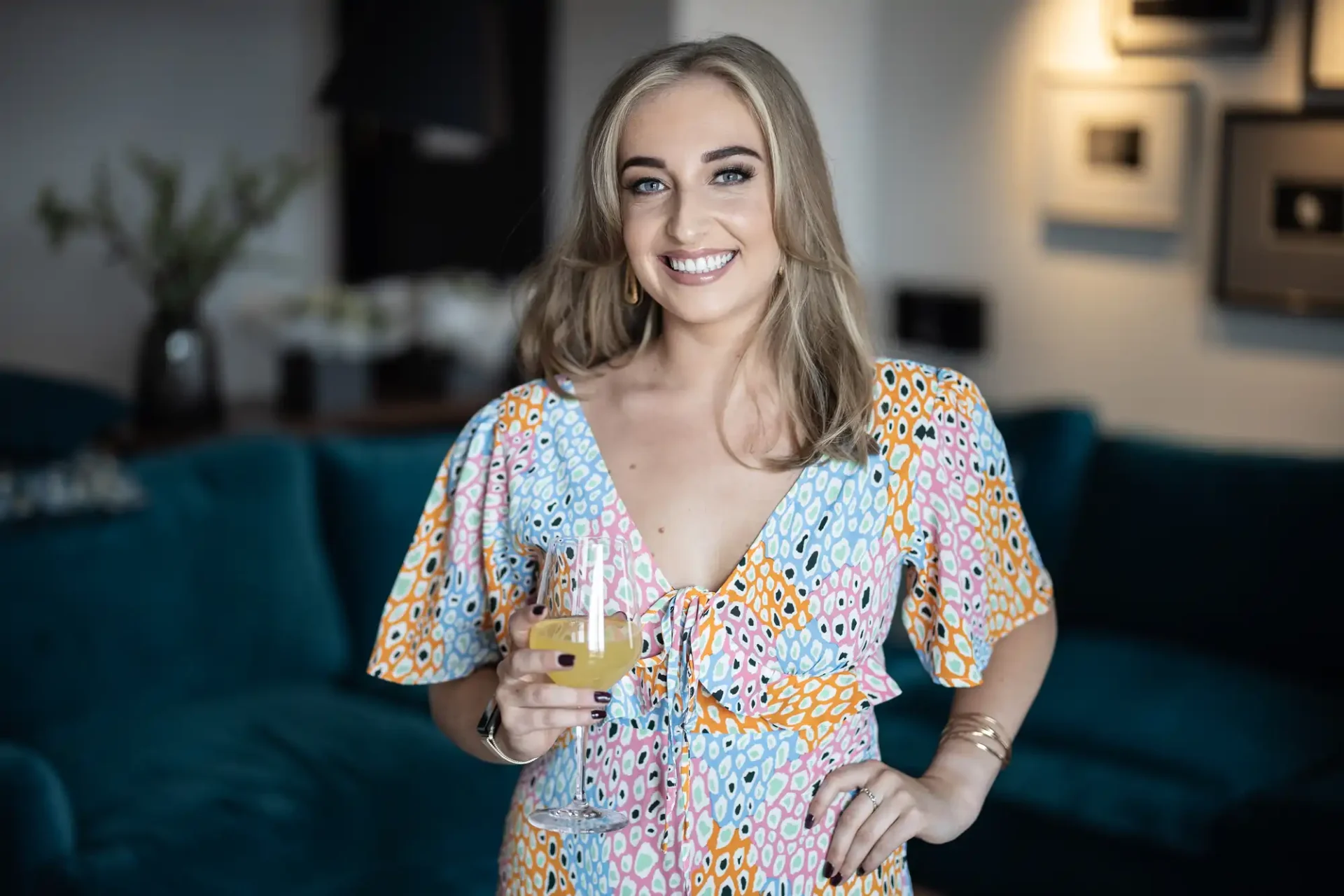 A smiling woman in a colorful dress holding a glass of wine, standing in a stylishly decorated room with blue sofas.