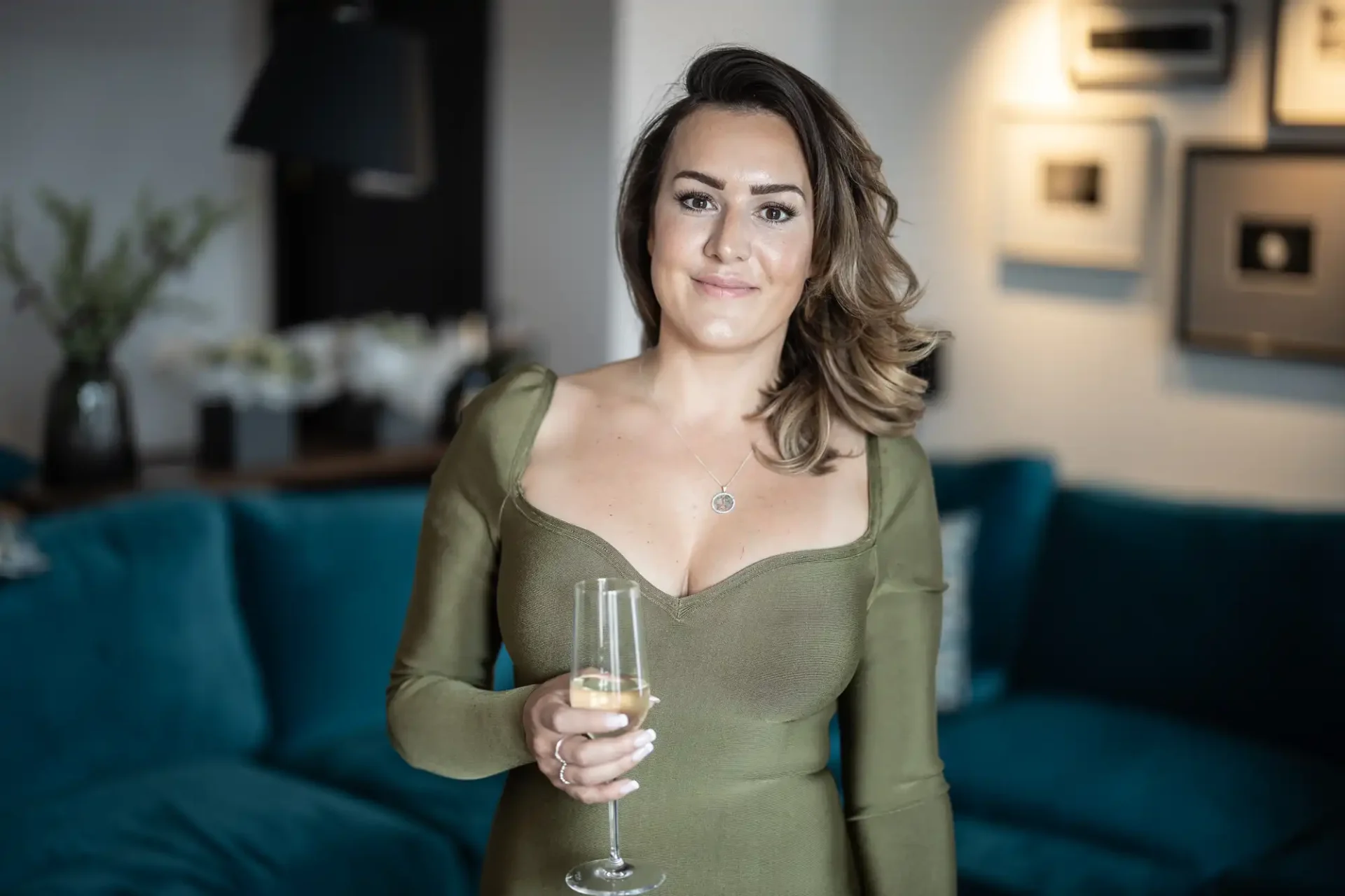 A woman in a green dress smiles while holding a glass of champagne, standing in a stylish living room.