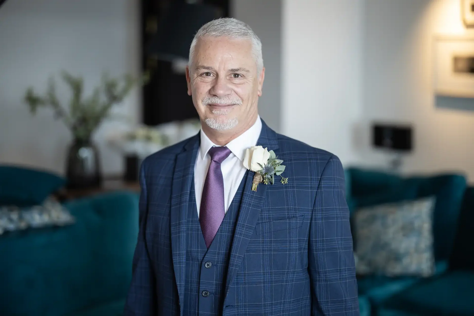 A mature man in a blue checkered suit and purple tie, smiling at a wedding, with a white boutonniere and blurred interior background.