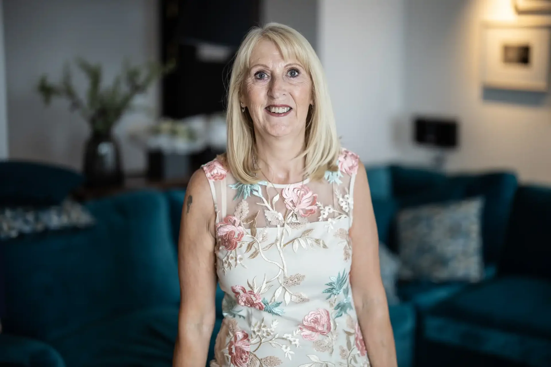 A smiling older woman with blonde hair wearing a floral dress standing in a room with blue sofas and a decorative background.