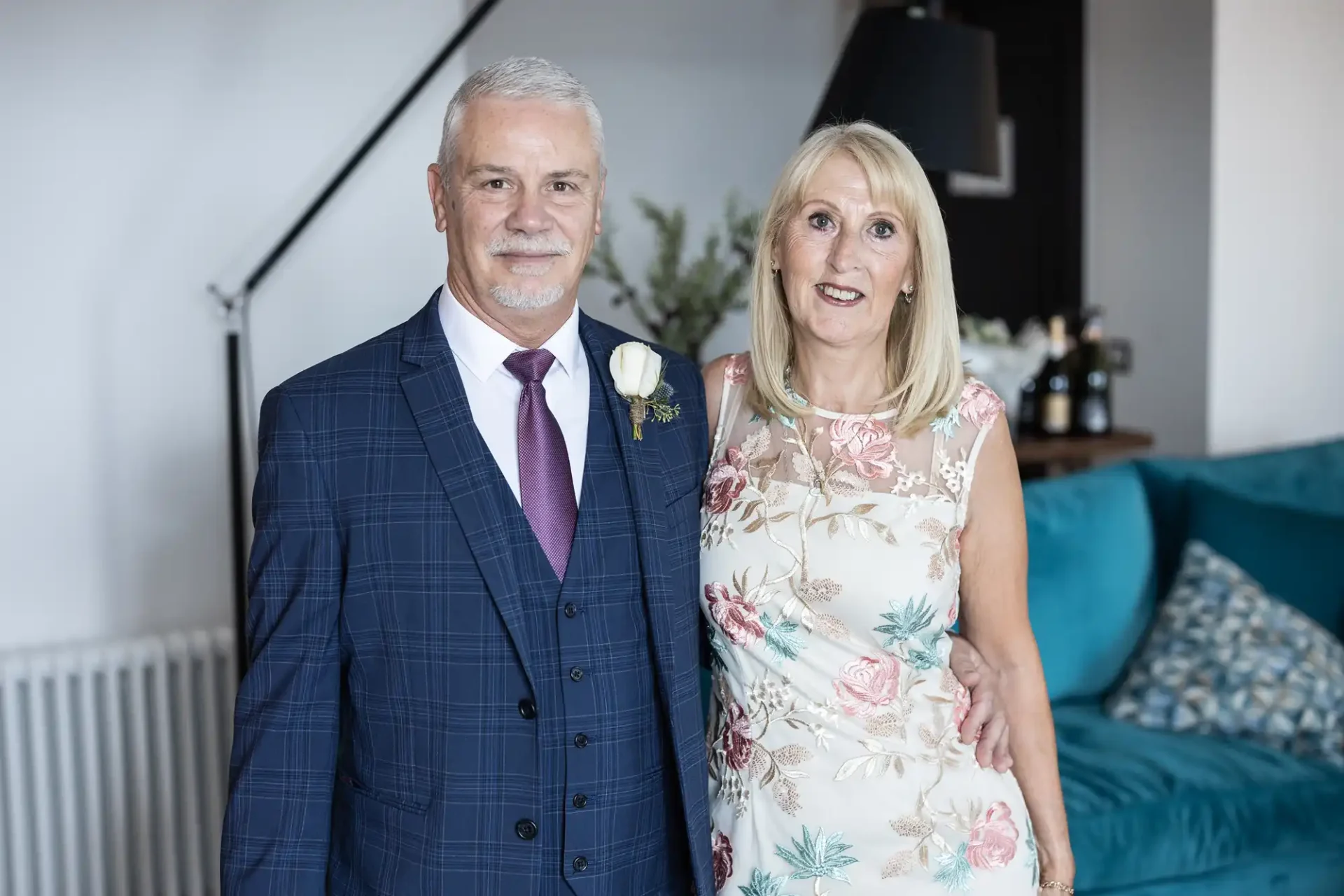 A mature couple dressed in formal attire—man in a blue checked suit and woman in a floral dress—smiling in a room with modern decor.