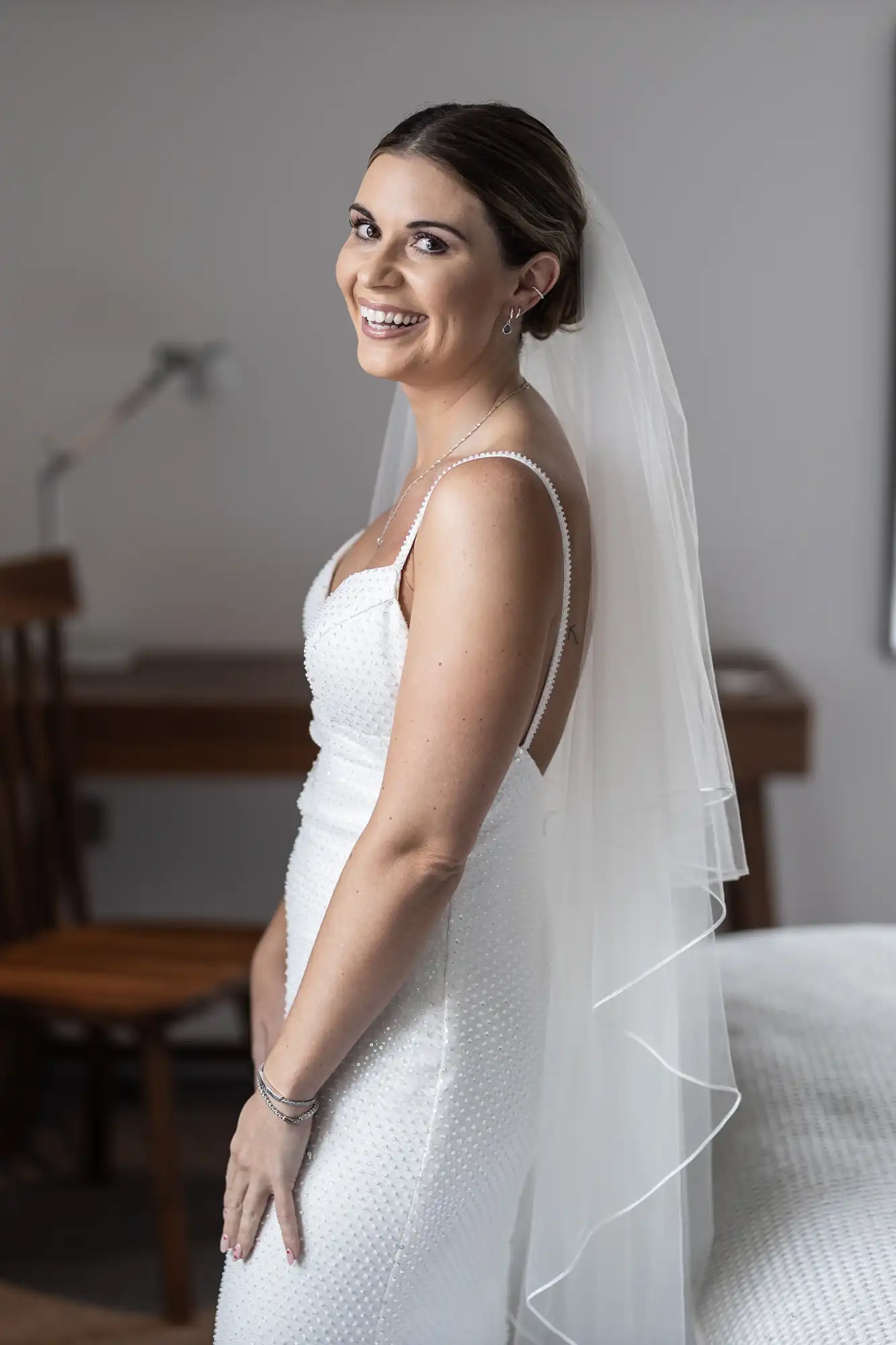 A smiling bride in a white beaded gown and veil stands in a room, turning to look over her shoulder.