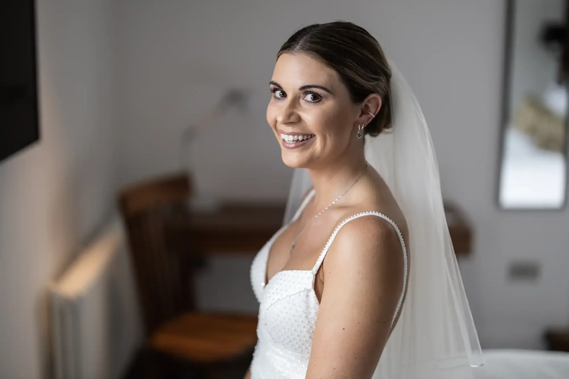 A bride in a white beaded wedding dress and veil smiling while looking over her shoulder in a softly lit room.