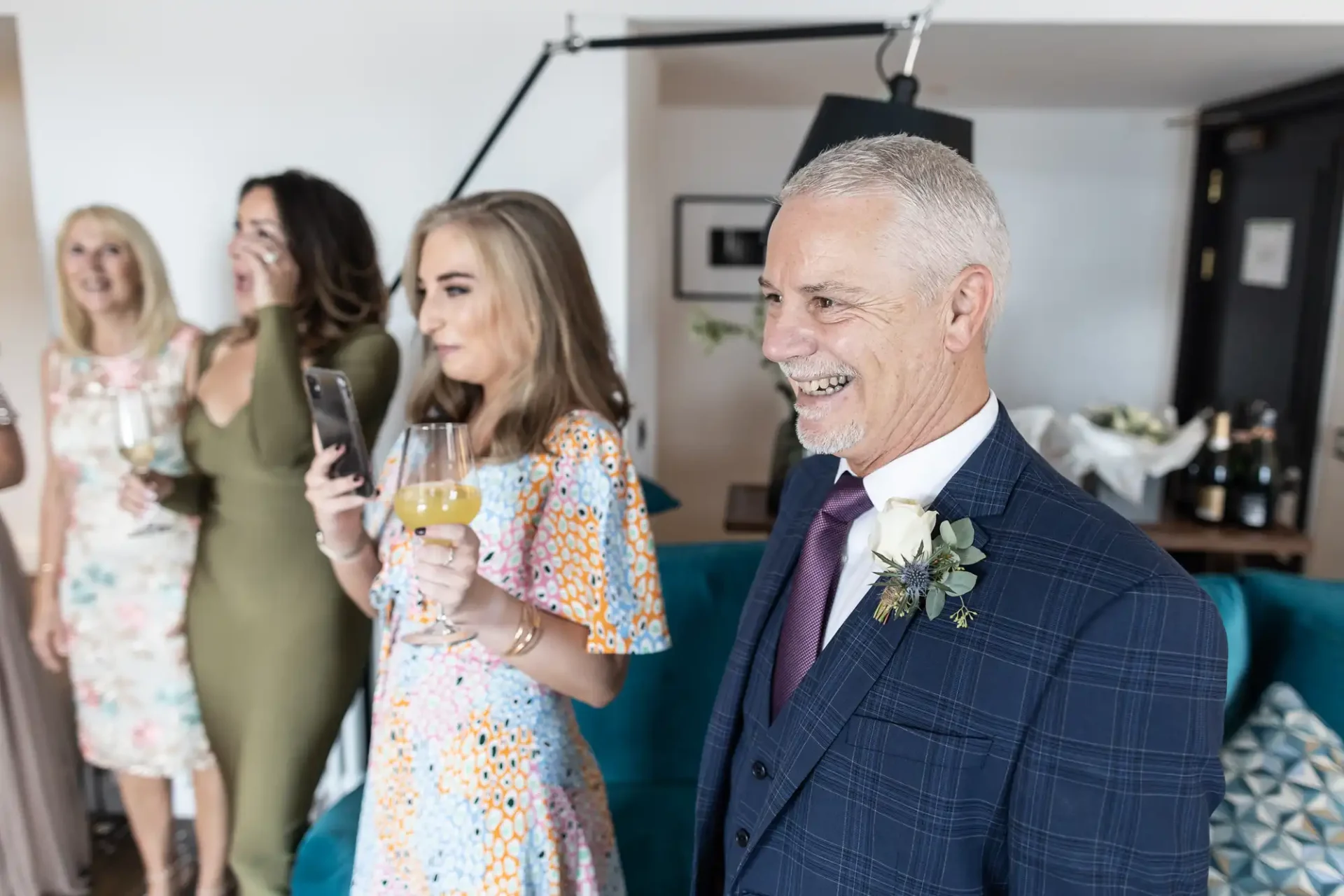 A smiling older man with a boutonniere stands in a room with three women holding drinks, who are engaged in conversation and laughter.