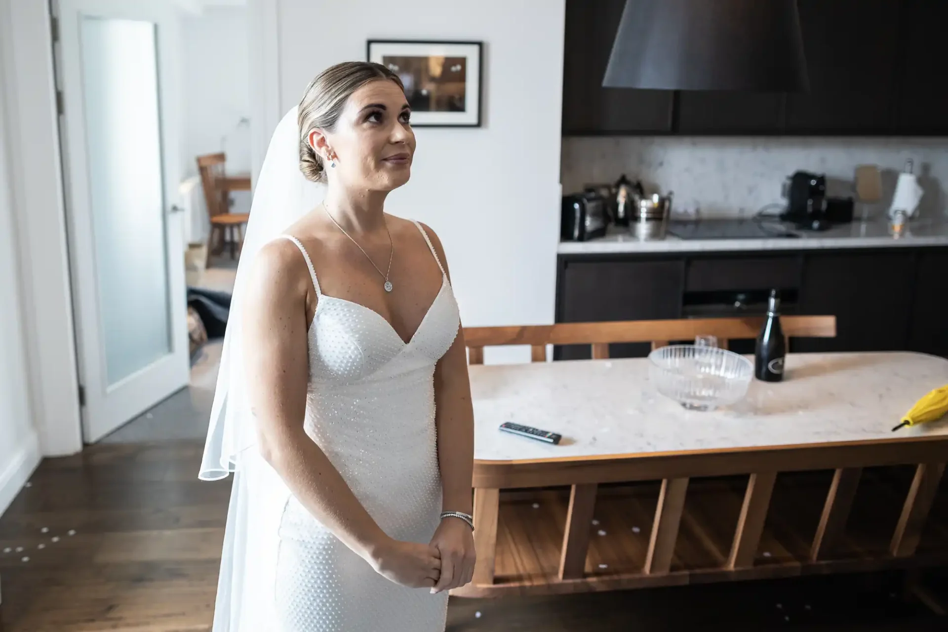 A bride in a white dress stands pensively in a modern kitchen, looking to the side.