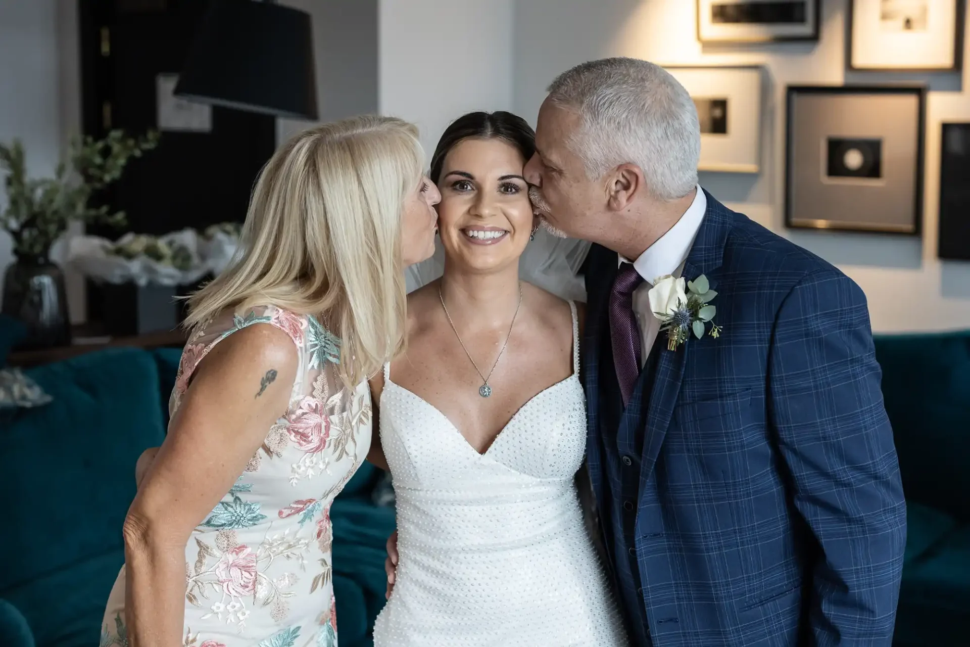 A bride smiling joyfully as her parents affectionately kiss her on each cheek in a decorated room.
