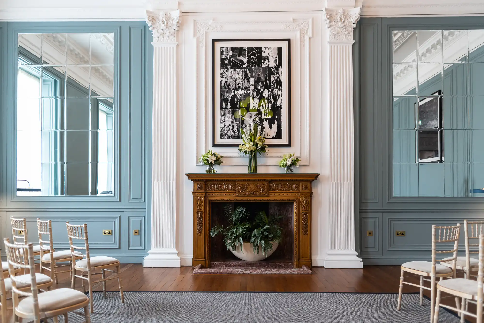 Elegant room with light blue walls, ornate golden fireplace with large abstract black and white artwork above, and neat rows of wooden chairs facing the fireplace.