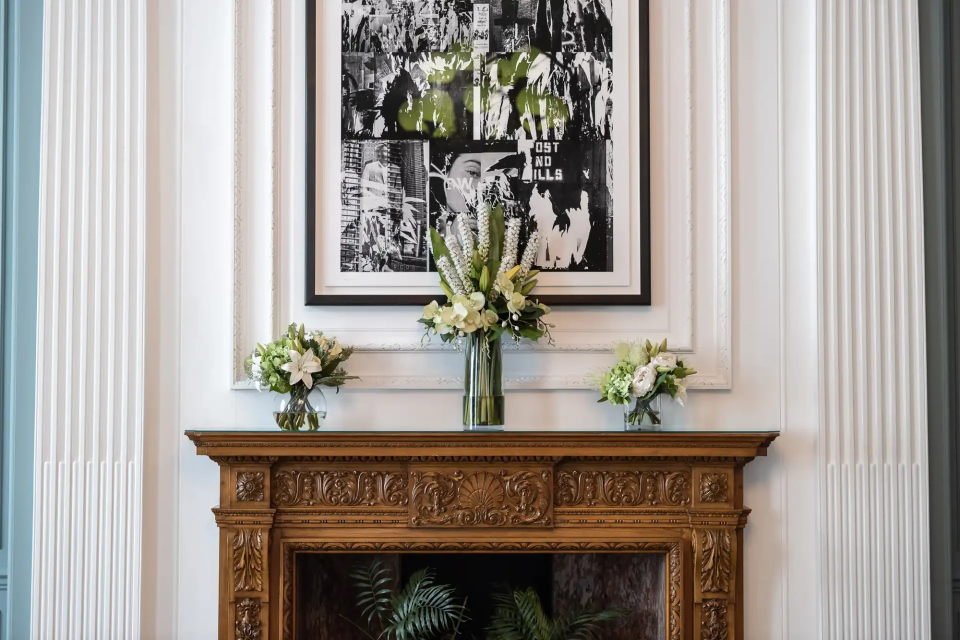 Elegant fireplace mantel with ornate wood carvings, flanked by white pillars and decorated with two floral arrangements and a vase of flowers, beneath a framed black and white poster.