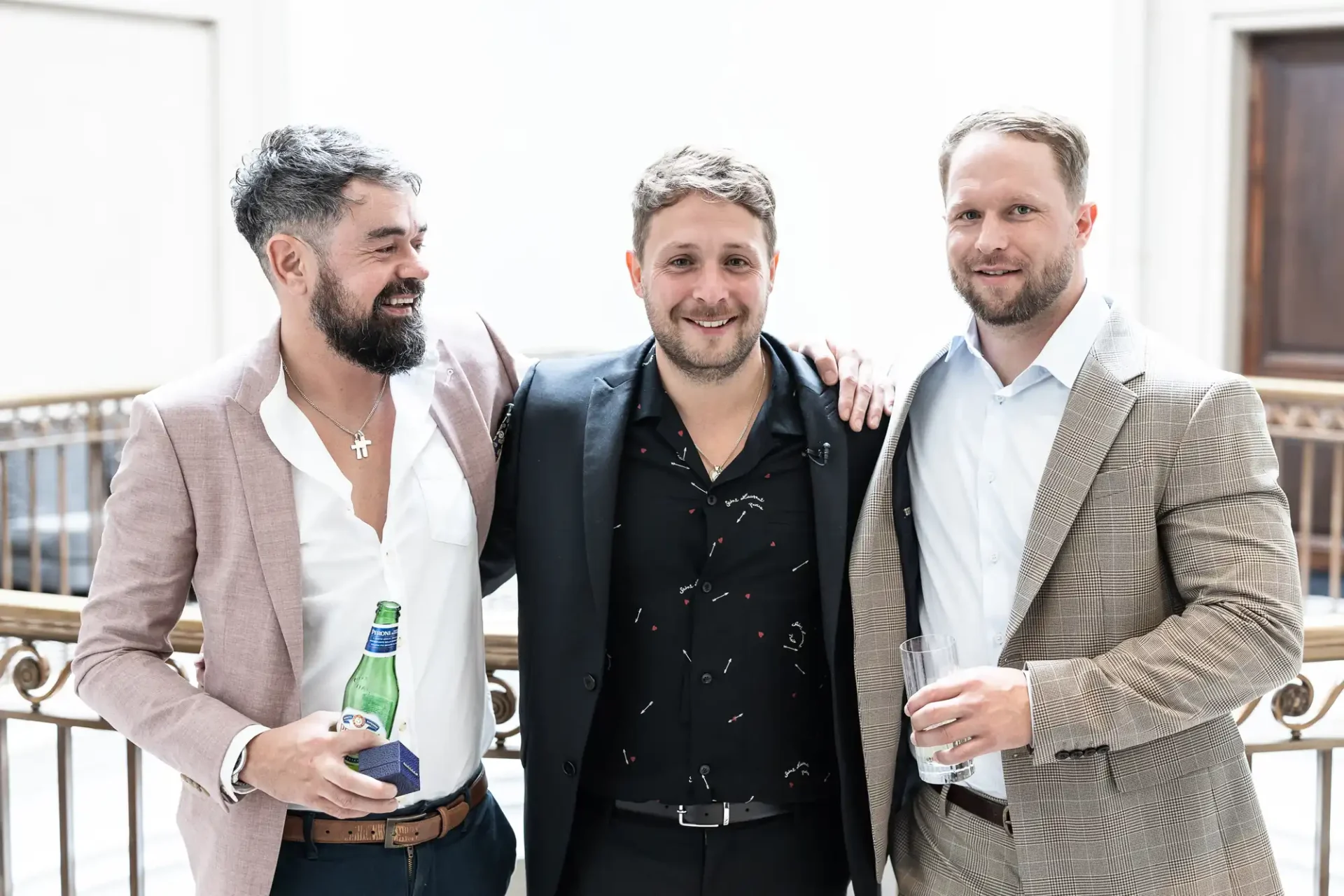 Three men in semi-formal attire smiling and posing together at an event, two holding drinks.