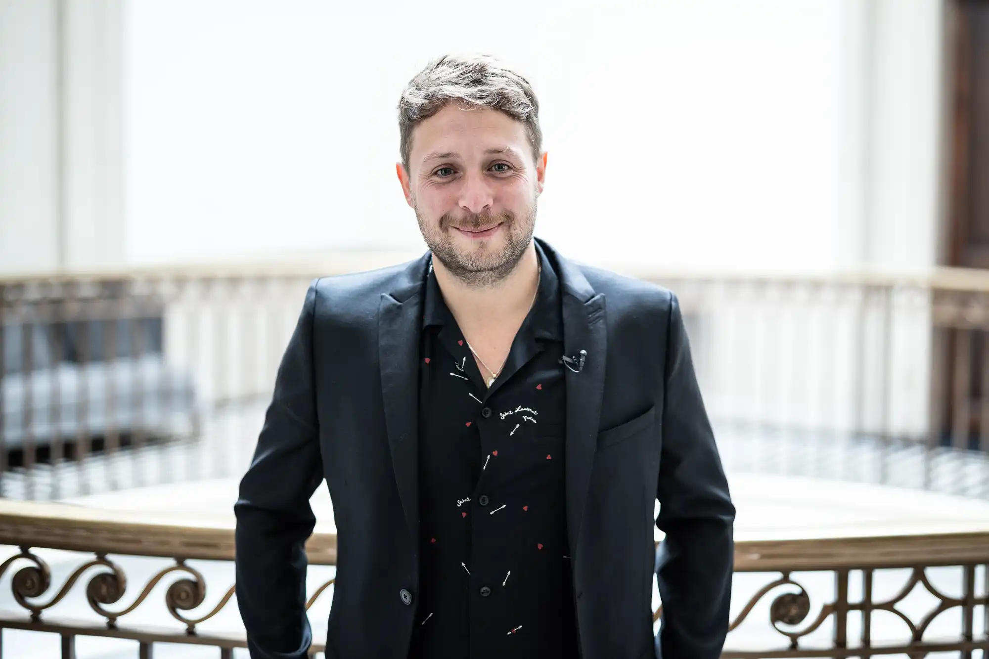 A man in a stylish black suit smiling at the camera, standing indoors with a white balustrade in the background.