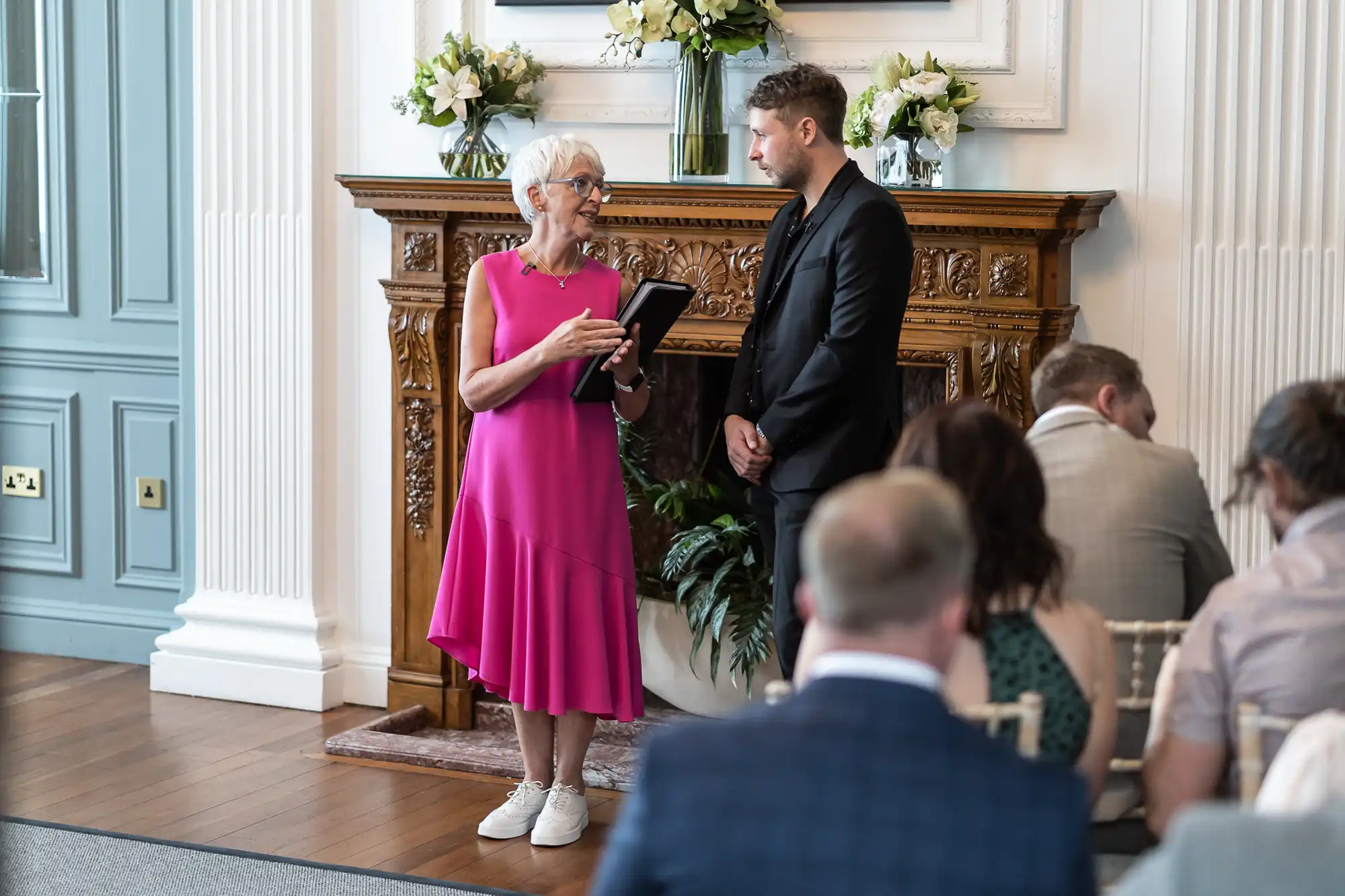 A woman in a pink dress holding a tablet stands beside a man in a suit, speaking to an audience in an elegant room with a fireplace and floral decorations.