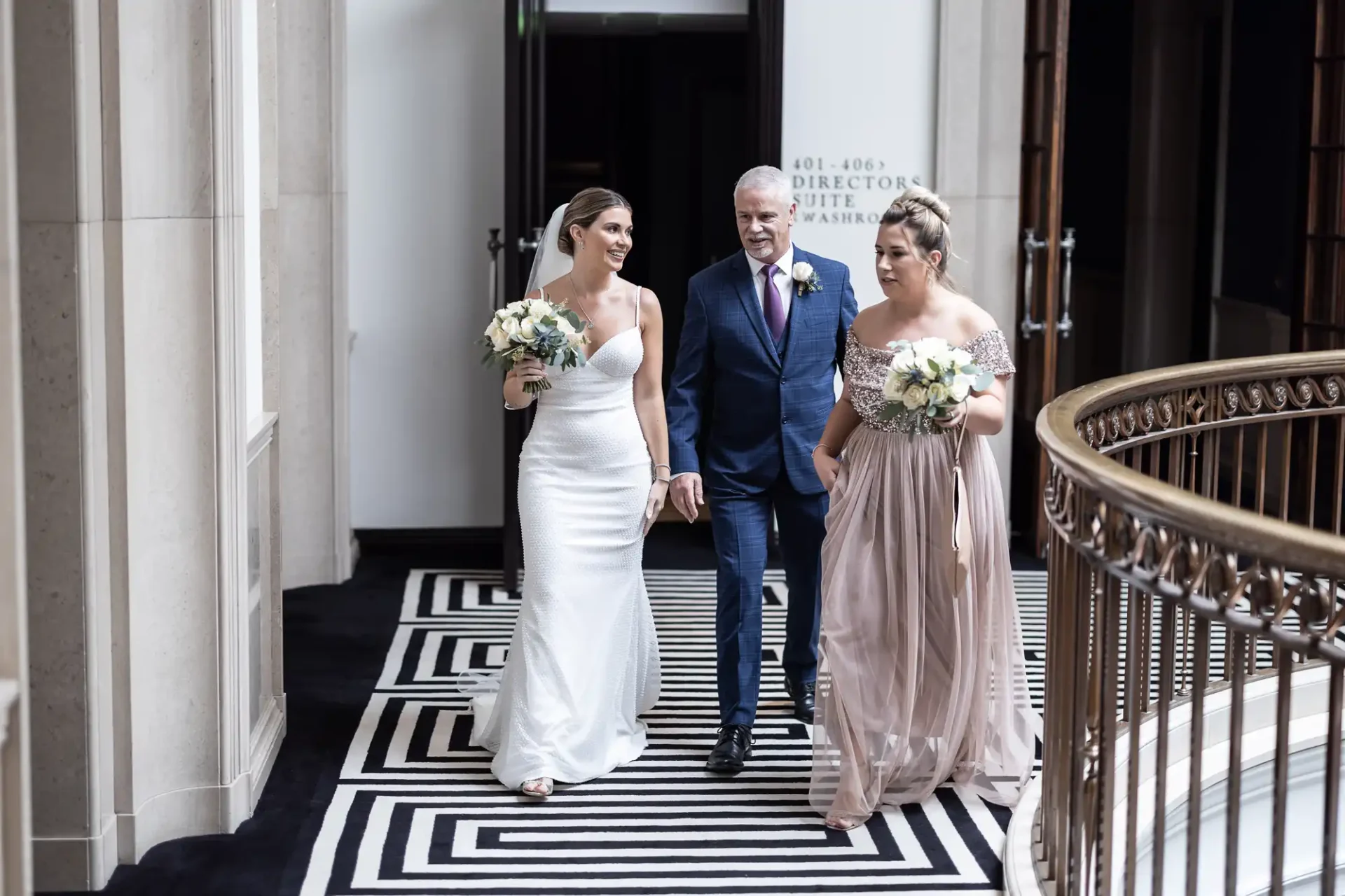 A bride in a white dress walks with her father and a bridesmaid in a beige dress down a corridor with a geometric carpet design.