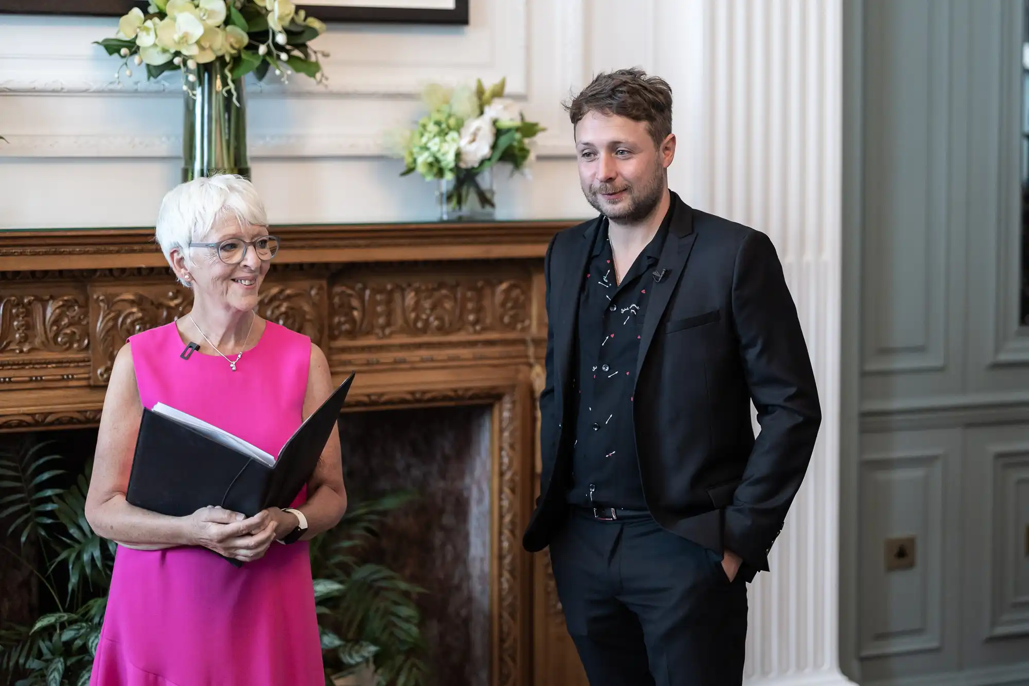 A woman in a pink dress holding a book stands beside a man in a black suit inside an elegant room with ornate decor.