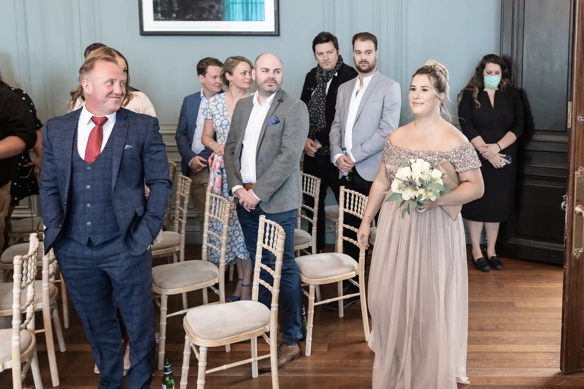 A bride walking down the aisle holding a bouquet, with an audience of guests watching, inside a room with vintage decor.