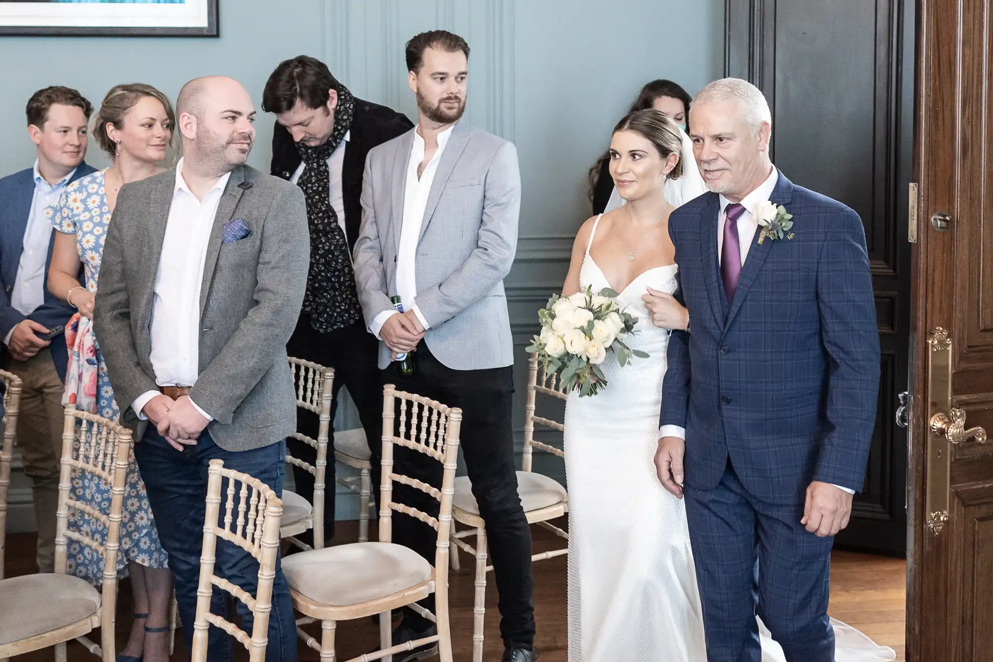 Bride walking down the aisle with her father, guests looking on, in an elegantly decorated room.