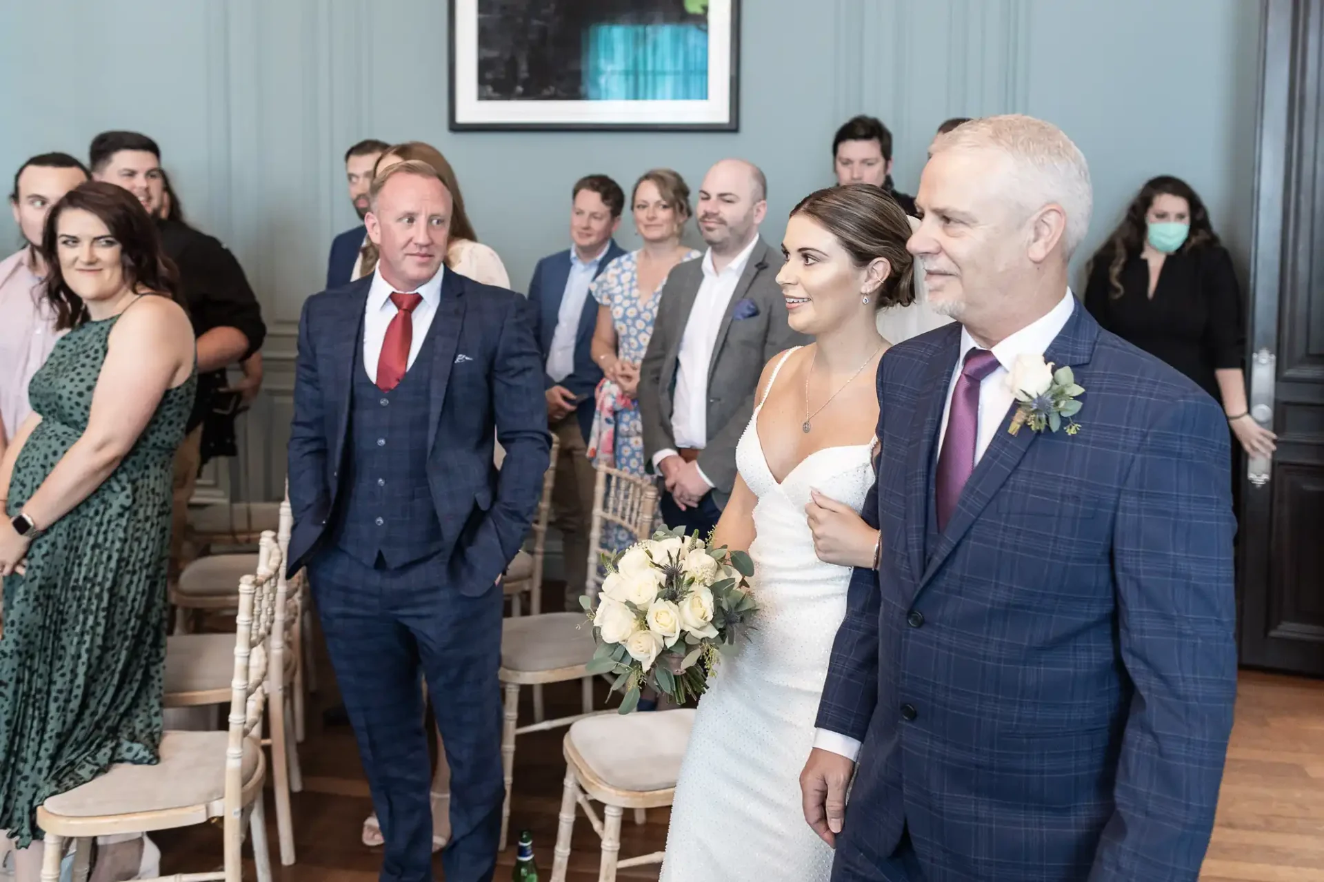 A bride walks down the aisle with an older man by her side, both smiling, as guests watch at an indoor wedding ceremony.