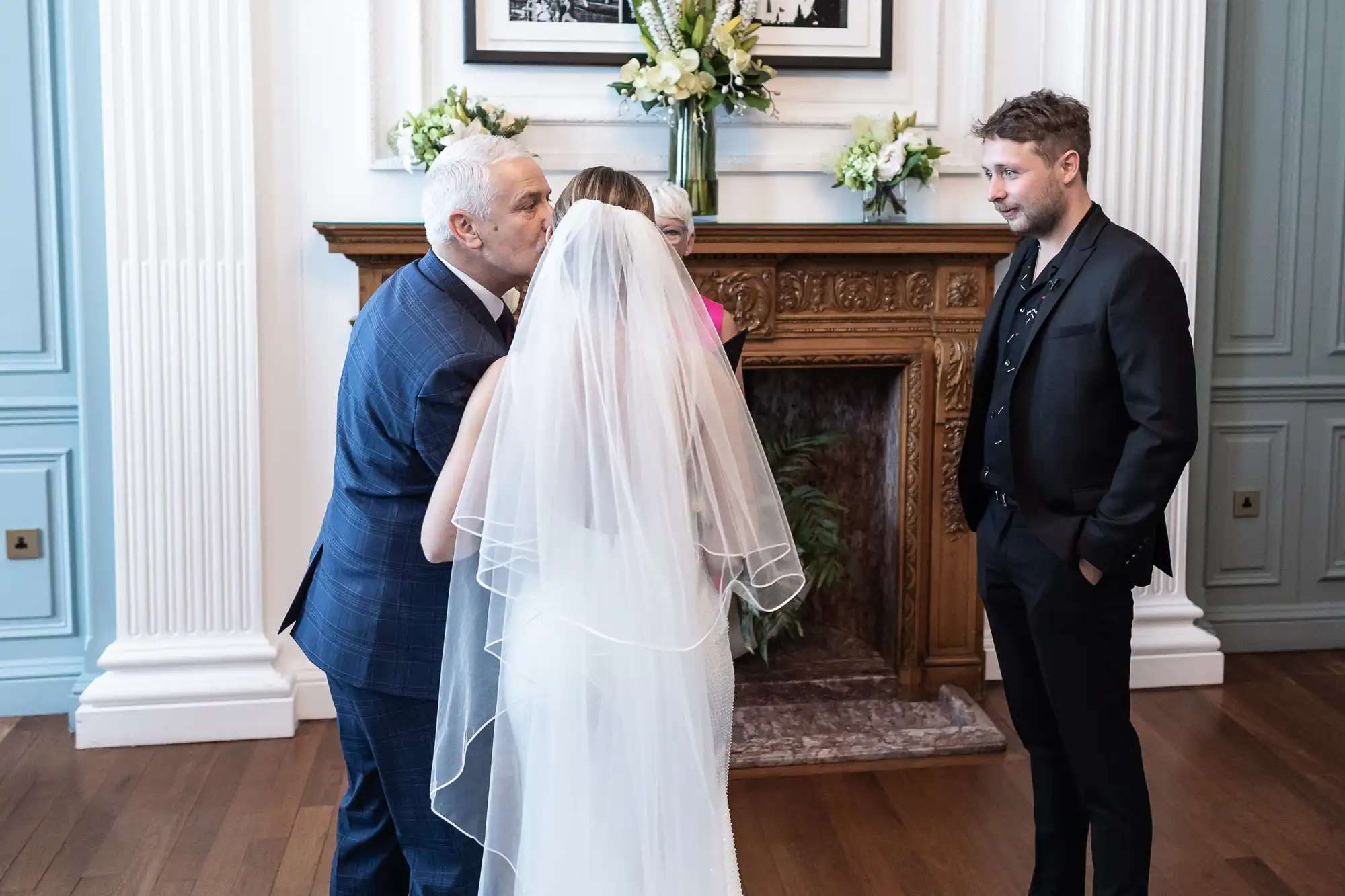 A father kisses his daughter on the forehead during her wedding ceremony, as the groom watches nearby in a room with elegant decor.