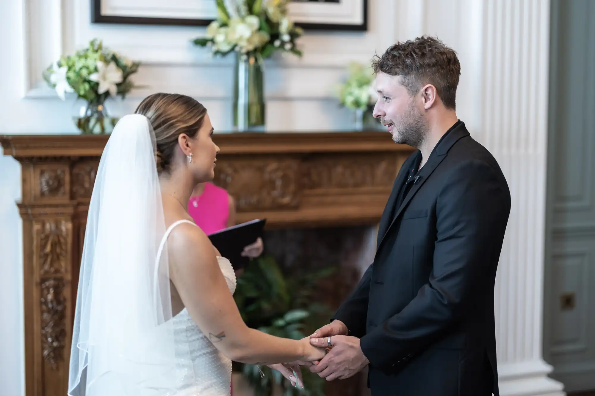 A bride and groom holding hands during a wedding ceremony in a room with a fireplace and elegant decor.