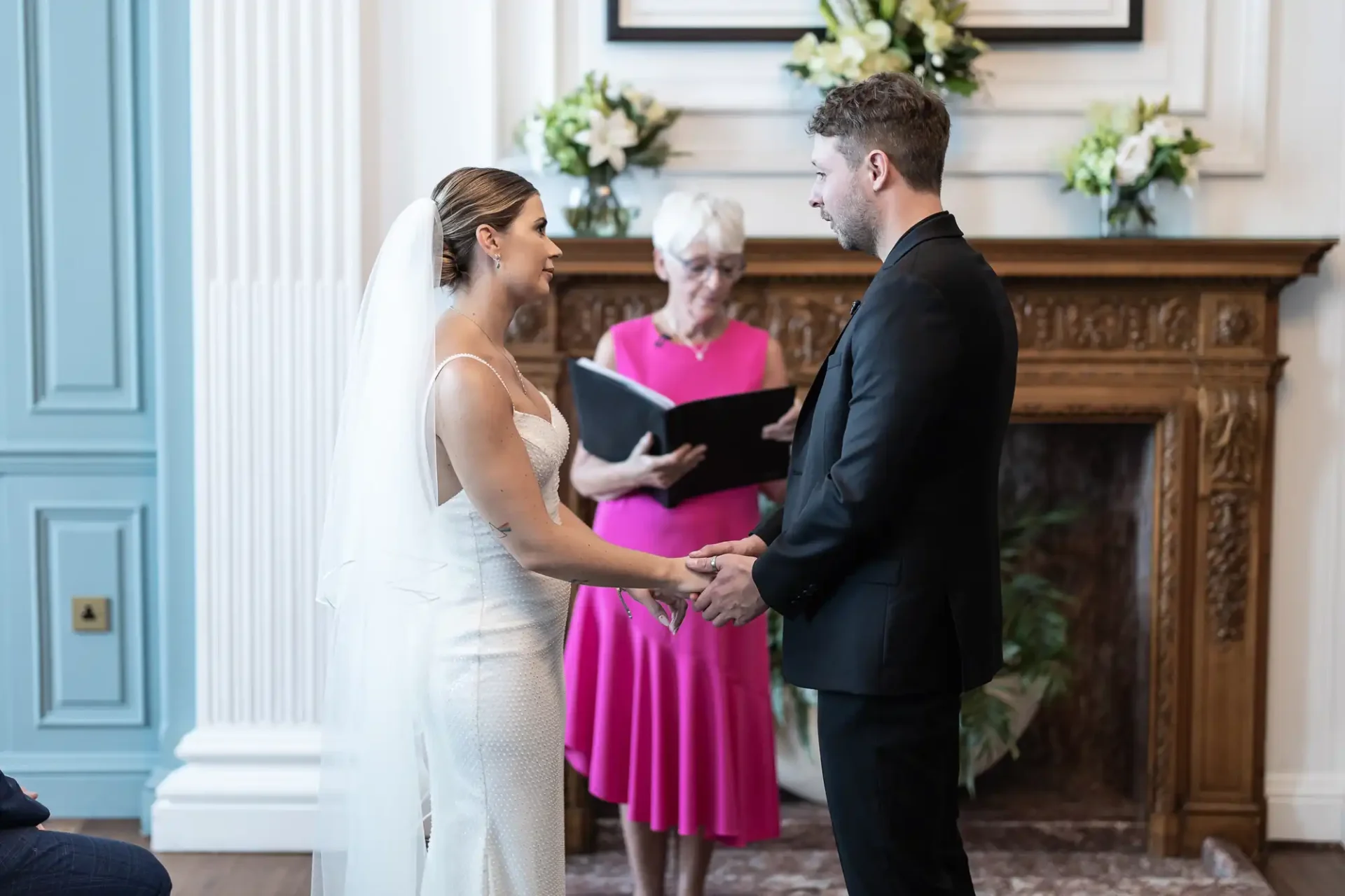 A bride and groom hold hands during their wedding ceremony, with an officiant in pink standing between them in a room with elegant decor.