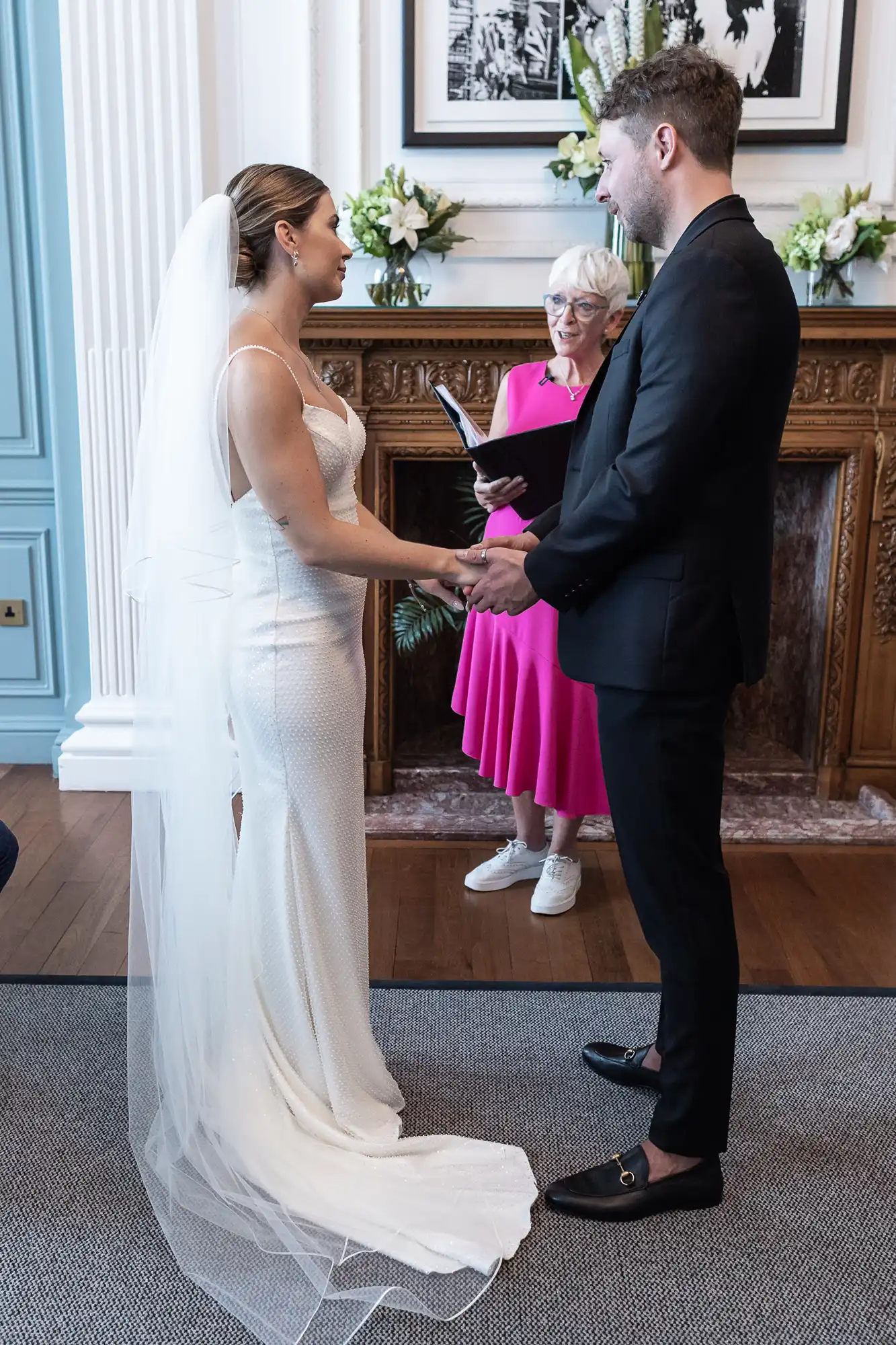 A bride in a white dress and veil and a groom in a dark suit hold hands during their wedding ceremony, with an officiant in pink standing by.