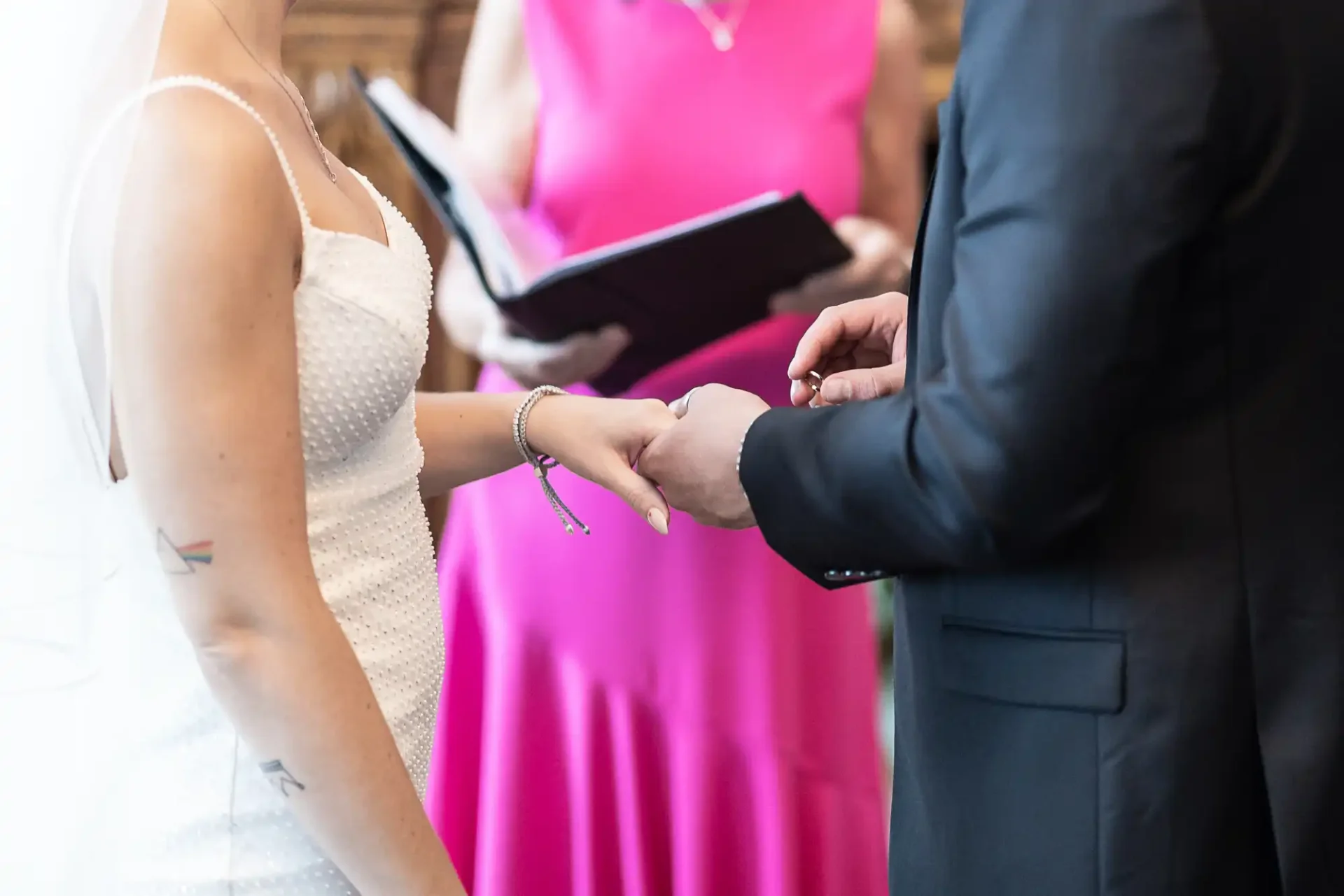 Bride in a white dress and groom in a black suit exchange rings during a wedding ceremony, with an officiant and bridesmaid in pink visible in the background.