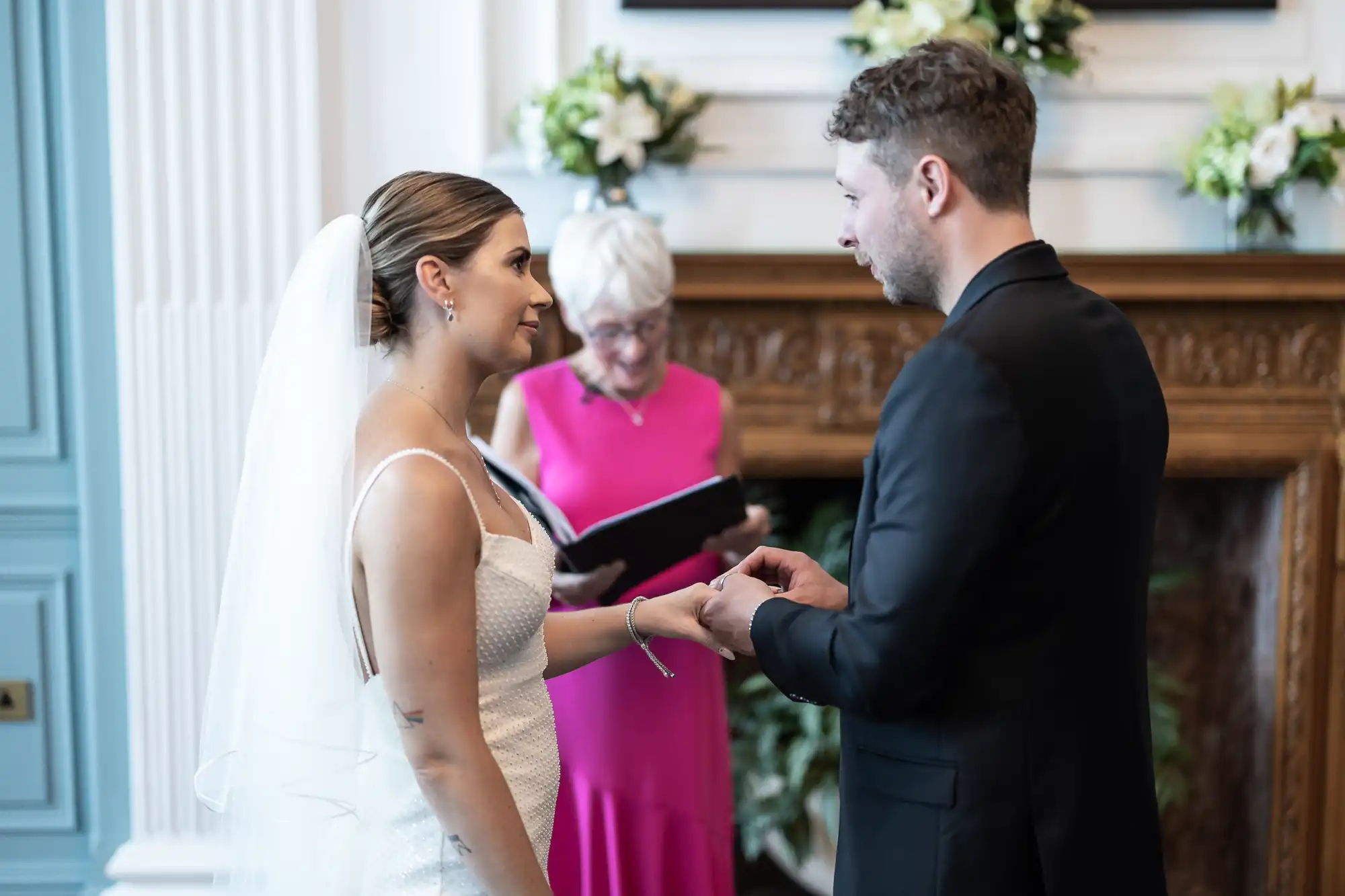 Bride and groom exchanging vows in front of an officiant at an indoor wedding ceremony, with elegant floral decorations in the background.