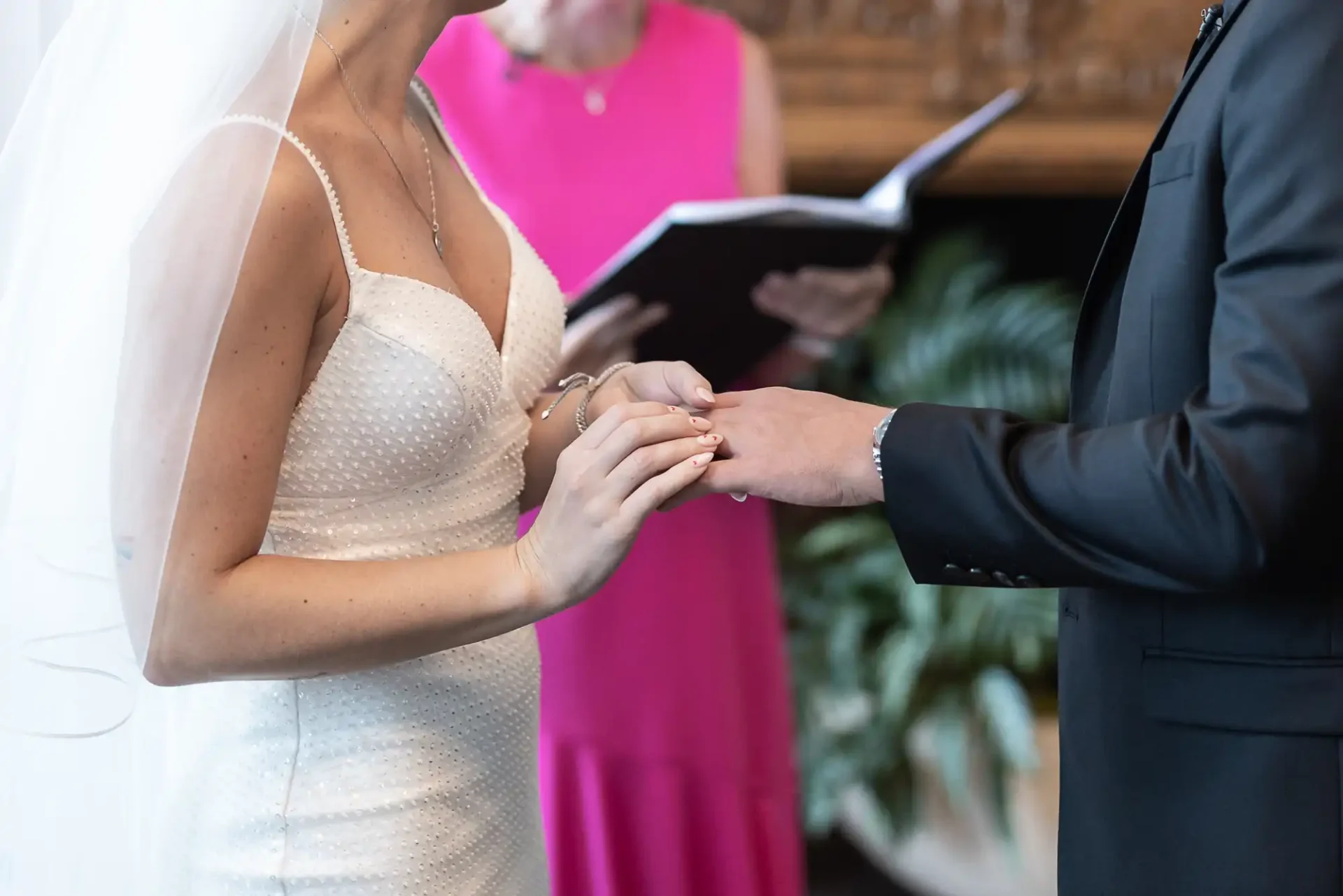 A bride in a white beaded dress and veil and a groom in a black suit exchange rings during a wedding ceremony, with an officiant in pink visible in the background.